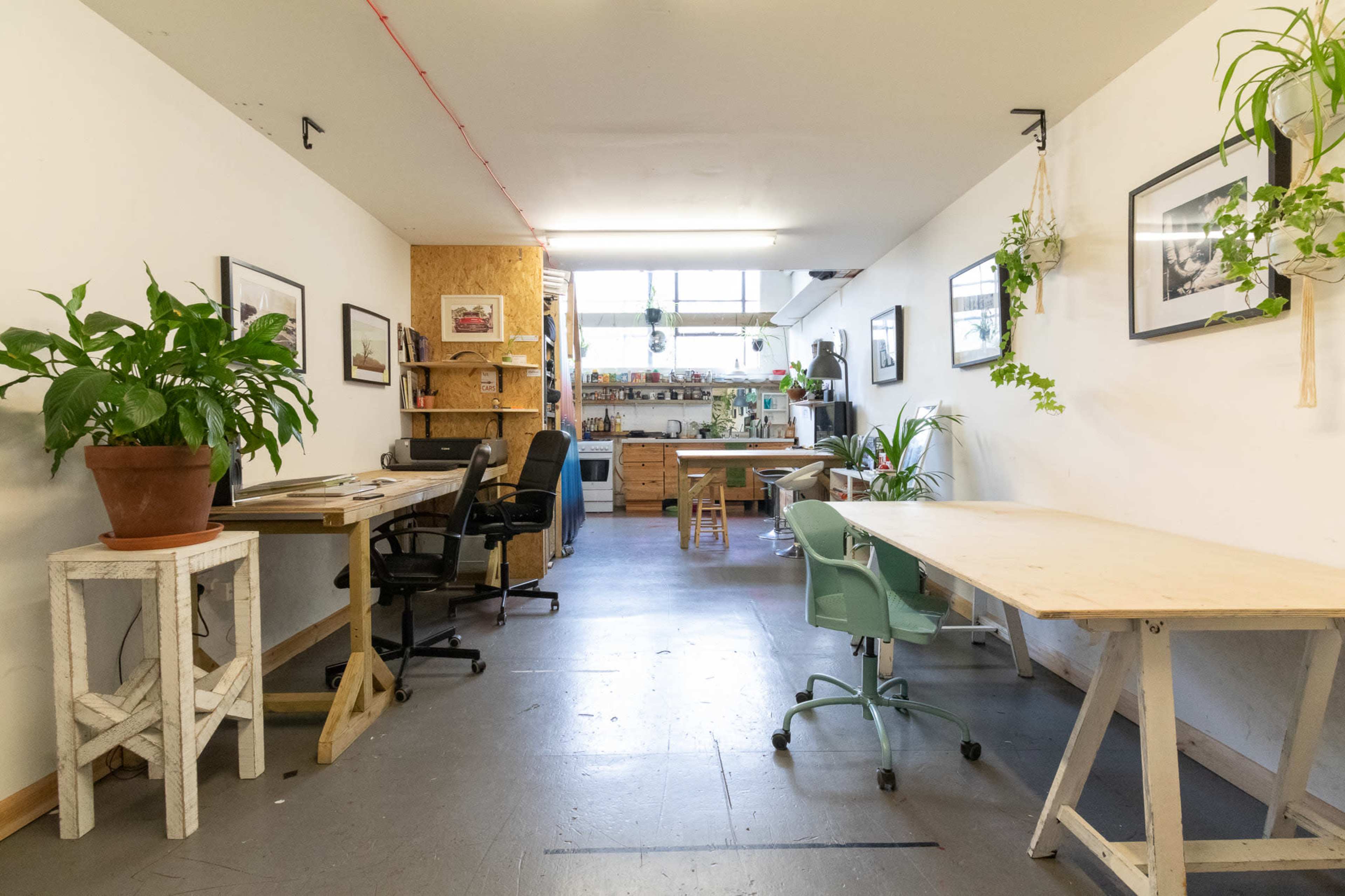 A spacious, well-lit artist studio featuring multiple work tables, chairs, and plants along the walls.