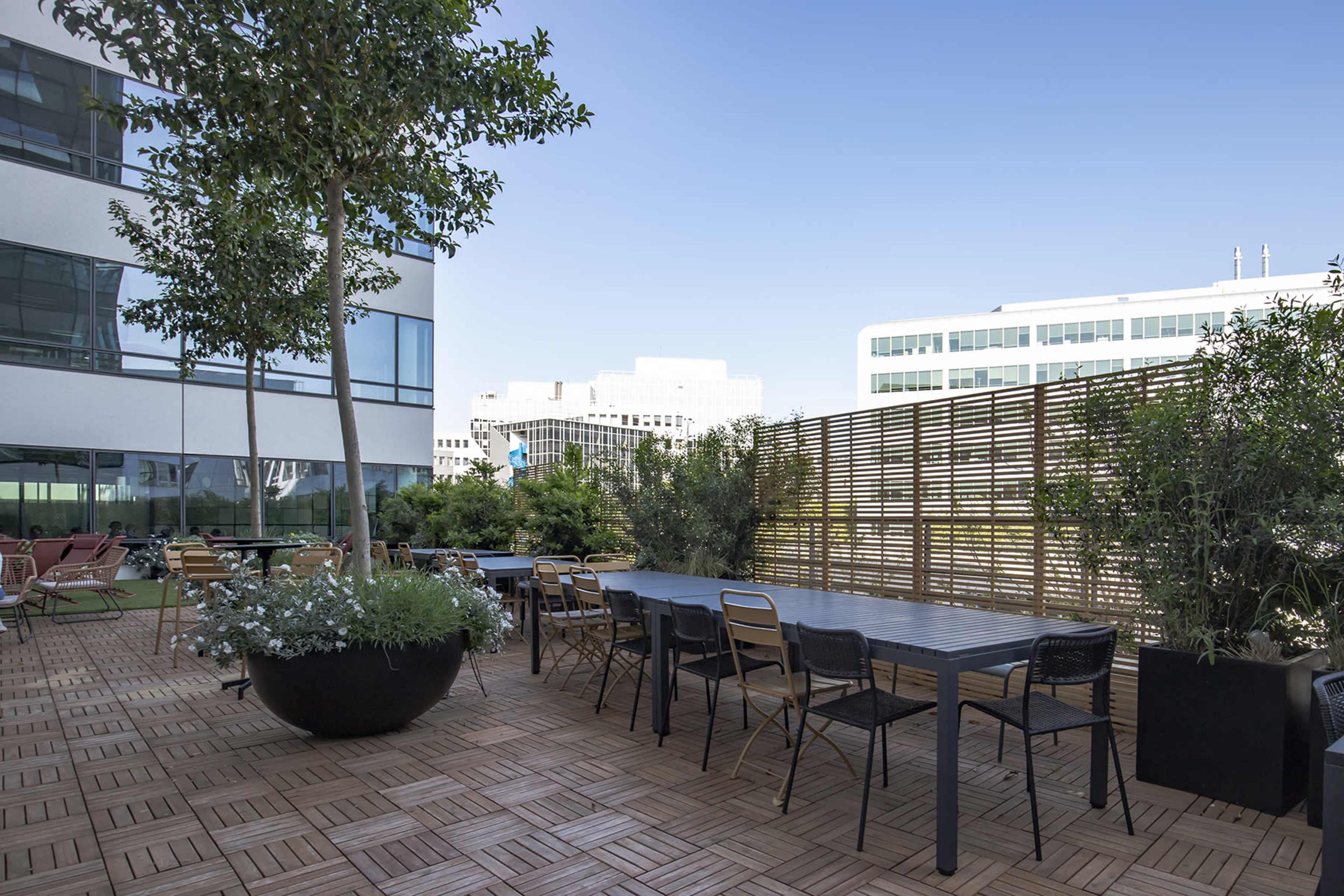 A rooftop terrace features a long table surrounded by chairs, with potted plants and wooden decking under a clear blue sky.