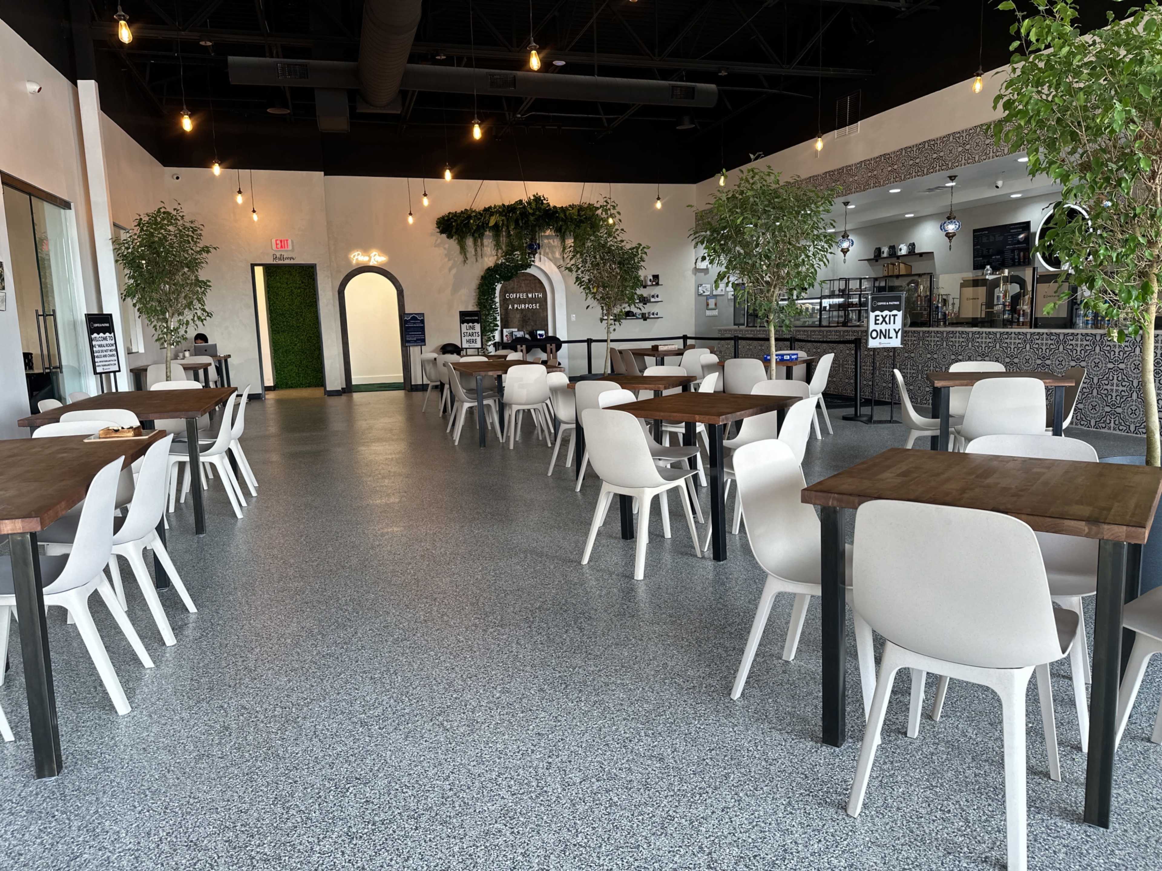 The image shows an empty dining area in a modern restaurant with wooden tables, white chairs, and indoor plants, illuminated by pendant lights.