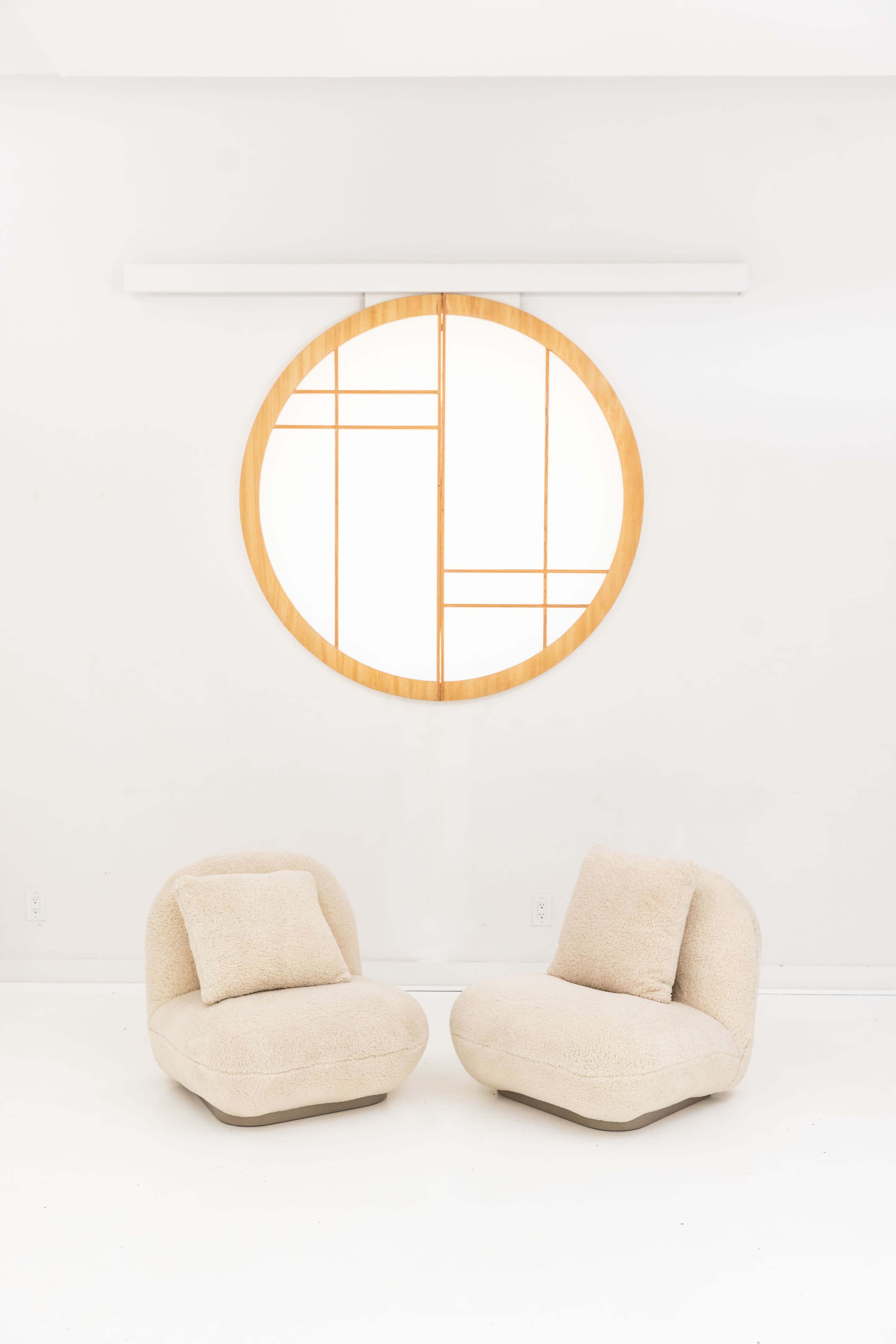 Two beige, rounded chairs positioned in front of a large circular wall mirror with a geometric wooden frame.