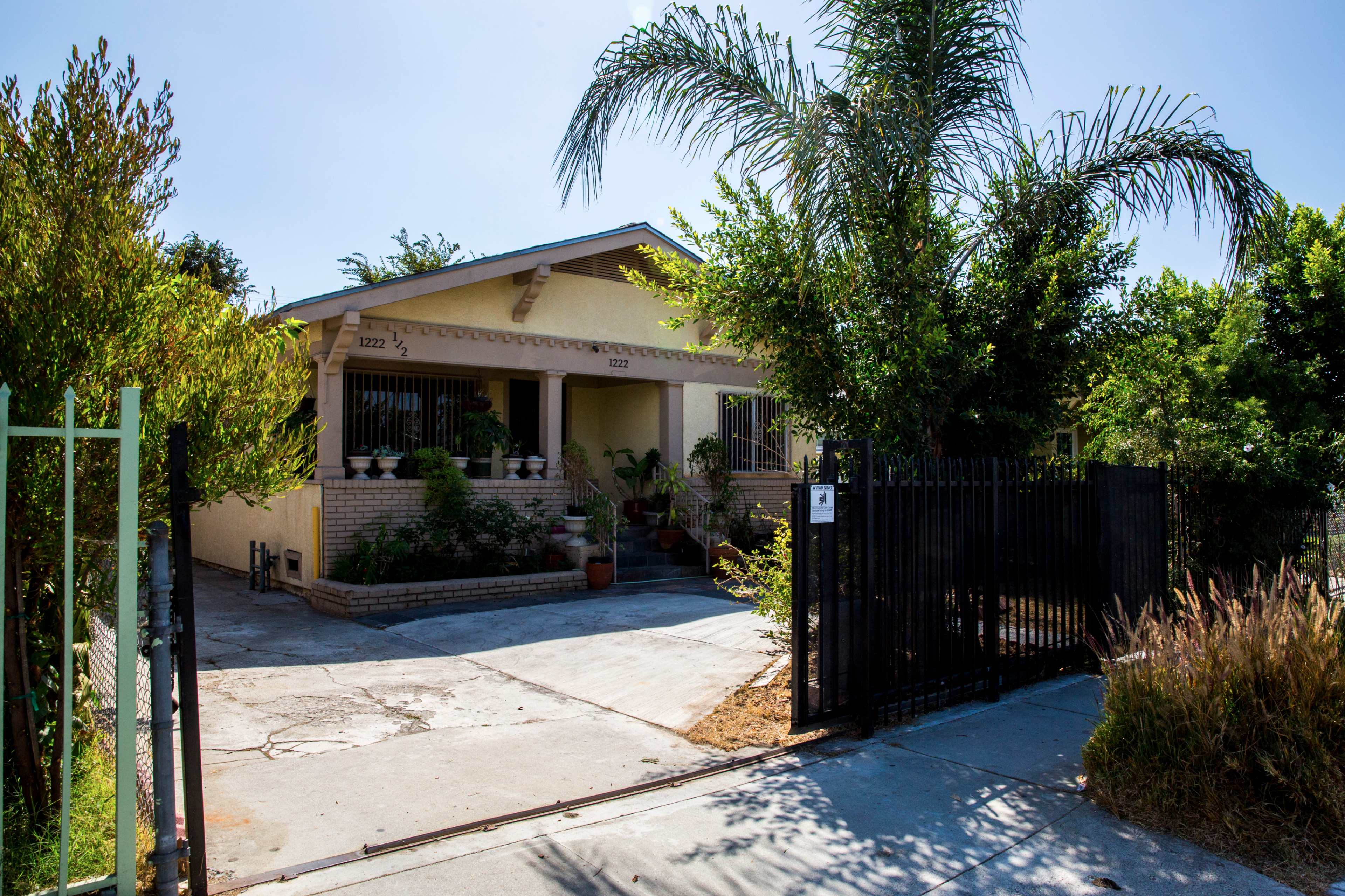 A single-story house with a red-tiled roof and a gated entrance is surrounded by trees and plants.