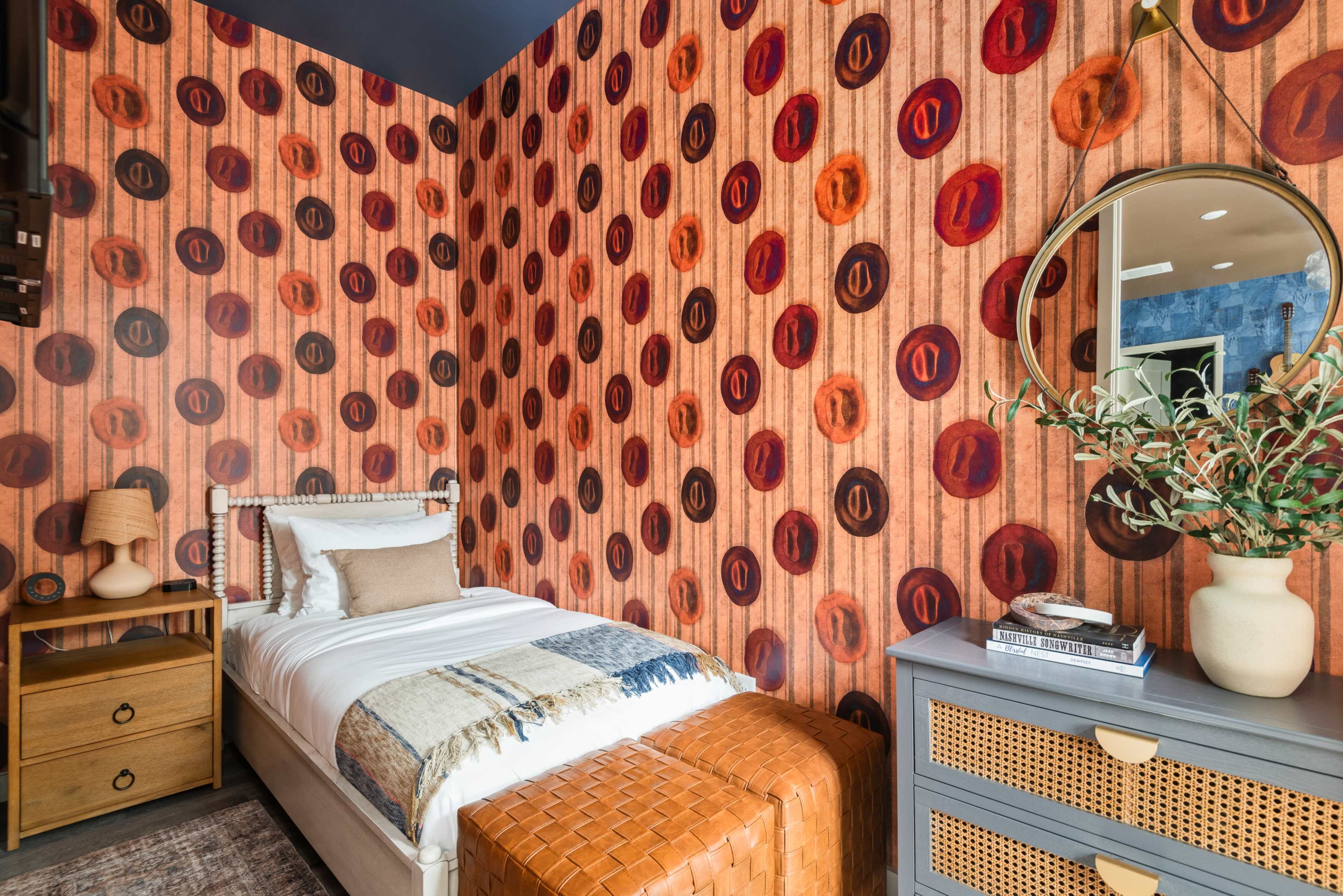 A small bedroom features a single bed with a decorative throw, a round mirror, and patterned wallpaper with circles against a textured background.