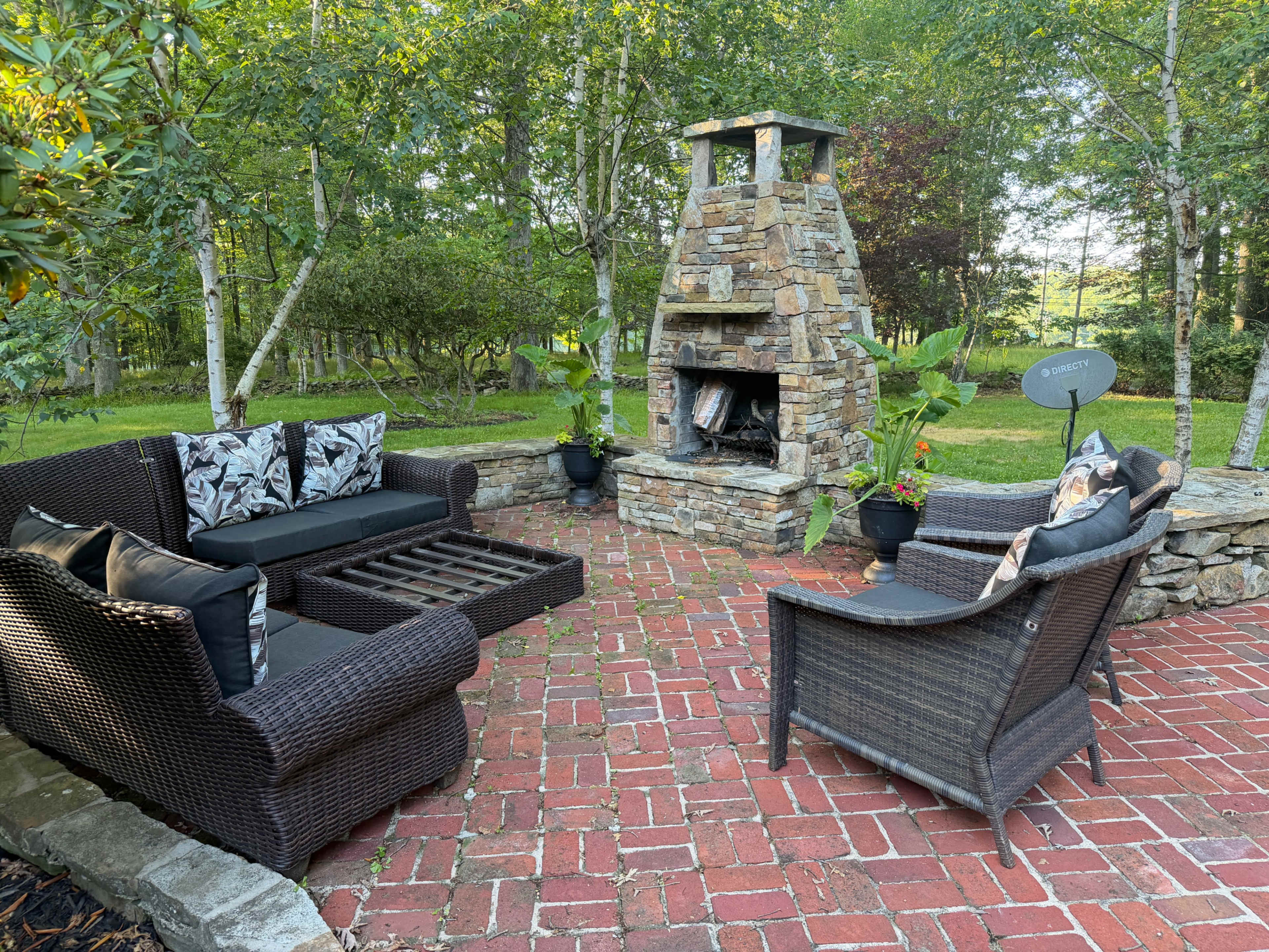 A cozy outdoor seating area features a stone fireplace, wicker furniture with cushions, and a brick patio surrounded by greenery.