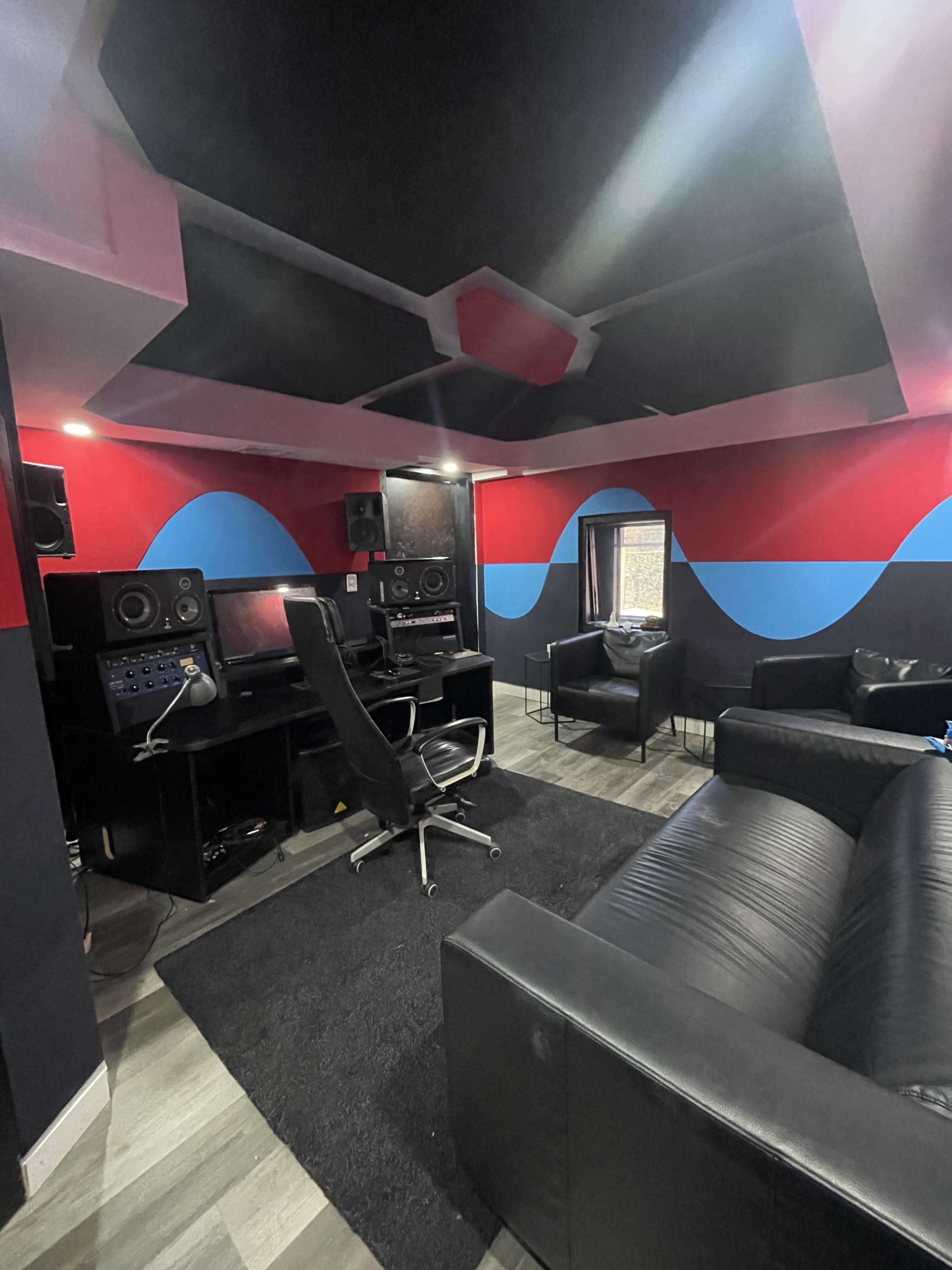 The image shows a small recording studio with black furniture, red and blue walls, soundproofing panels, and audio equipment.