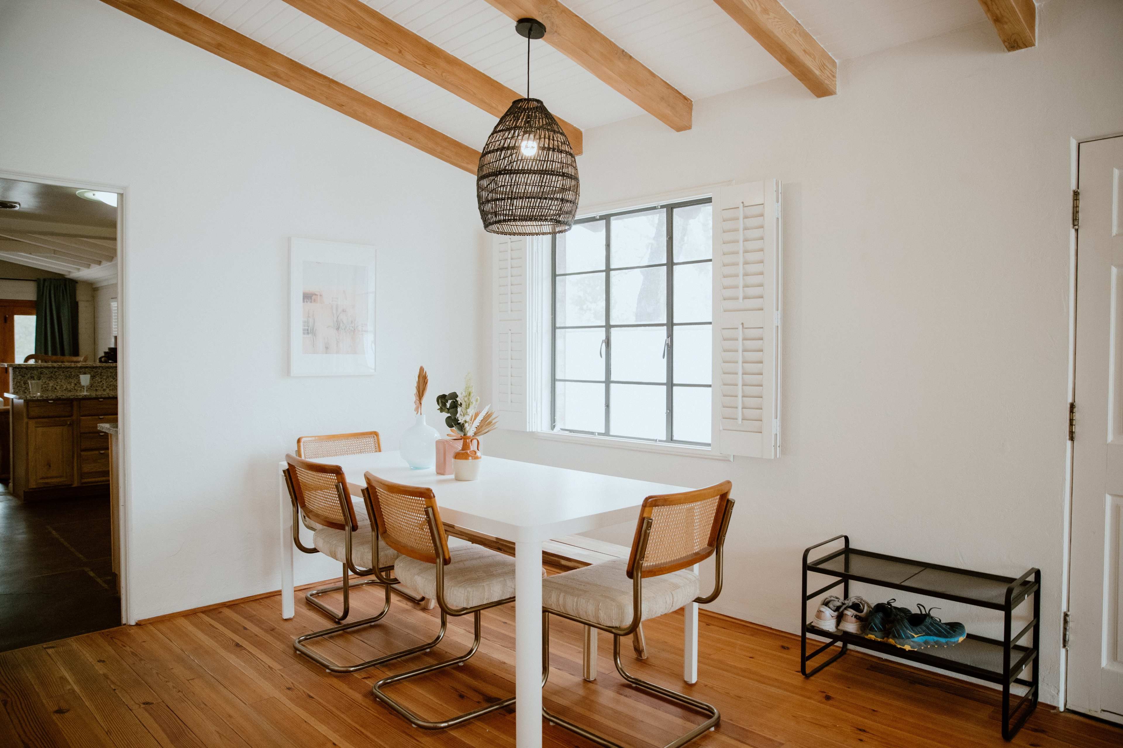 A clean dining area features a white table and four chairs, illuminated by a pendant lamp, with a window adorned with shutters and a small shoe rack nearby.