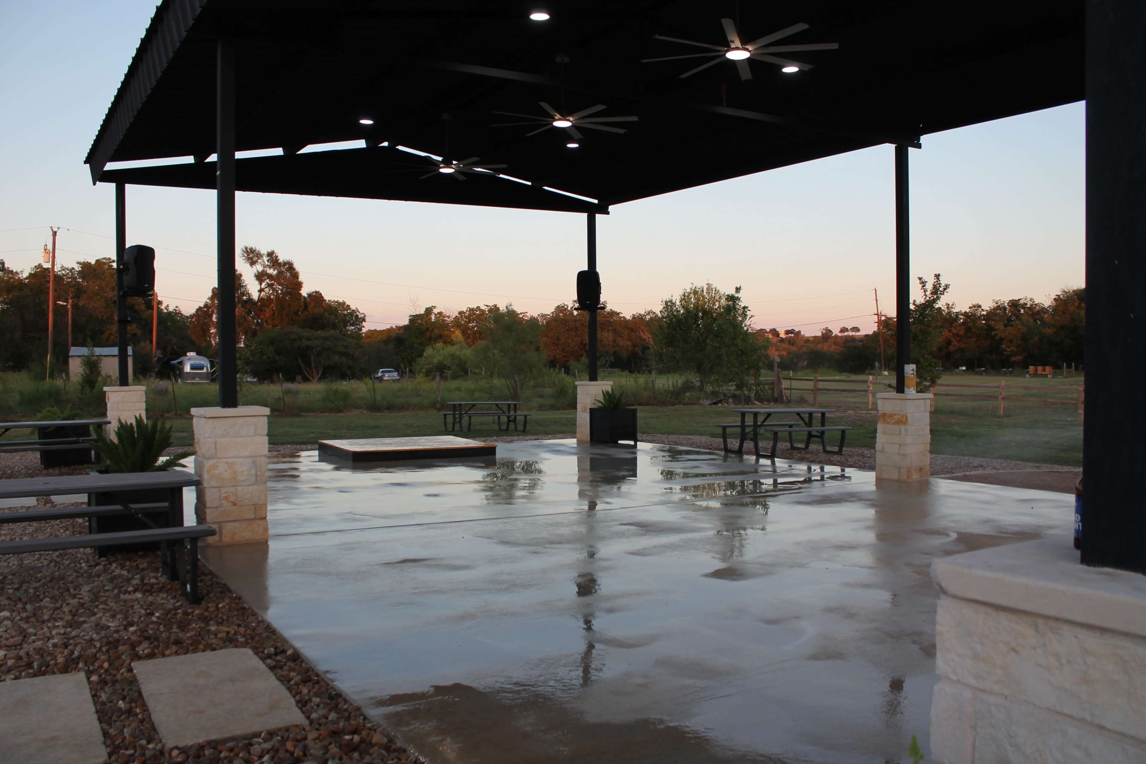 The image shows a covered outdoor pavilion with a polished concrete floor, picnic tables, and modern lighting fixtures, set in a grassy area with trees in the background.
