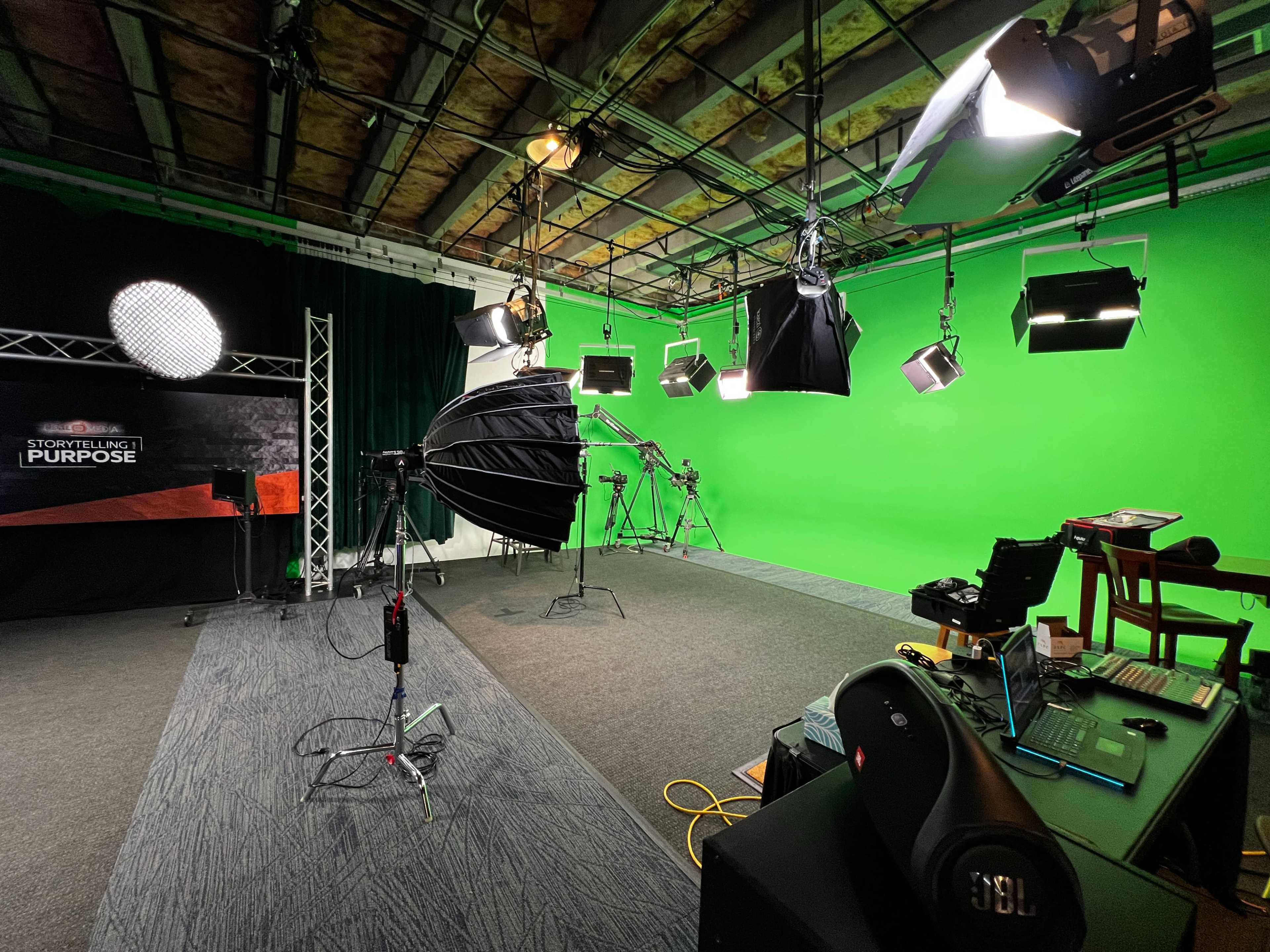 The image shows a green screen studio setup with various lighting equipment, cameras, and reflective umbrellas arranged on a textured floor.