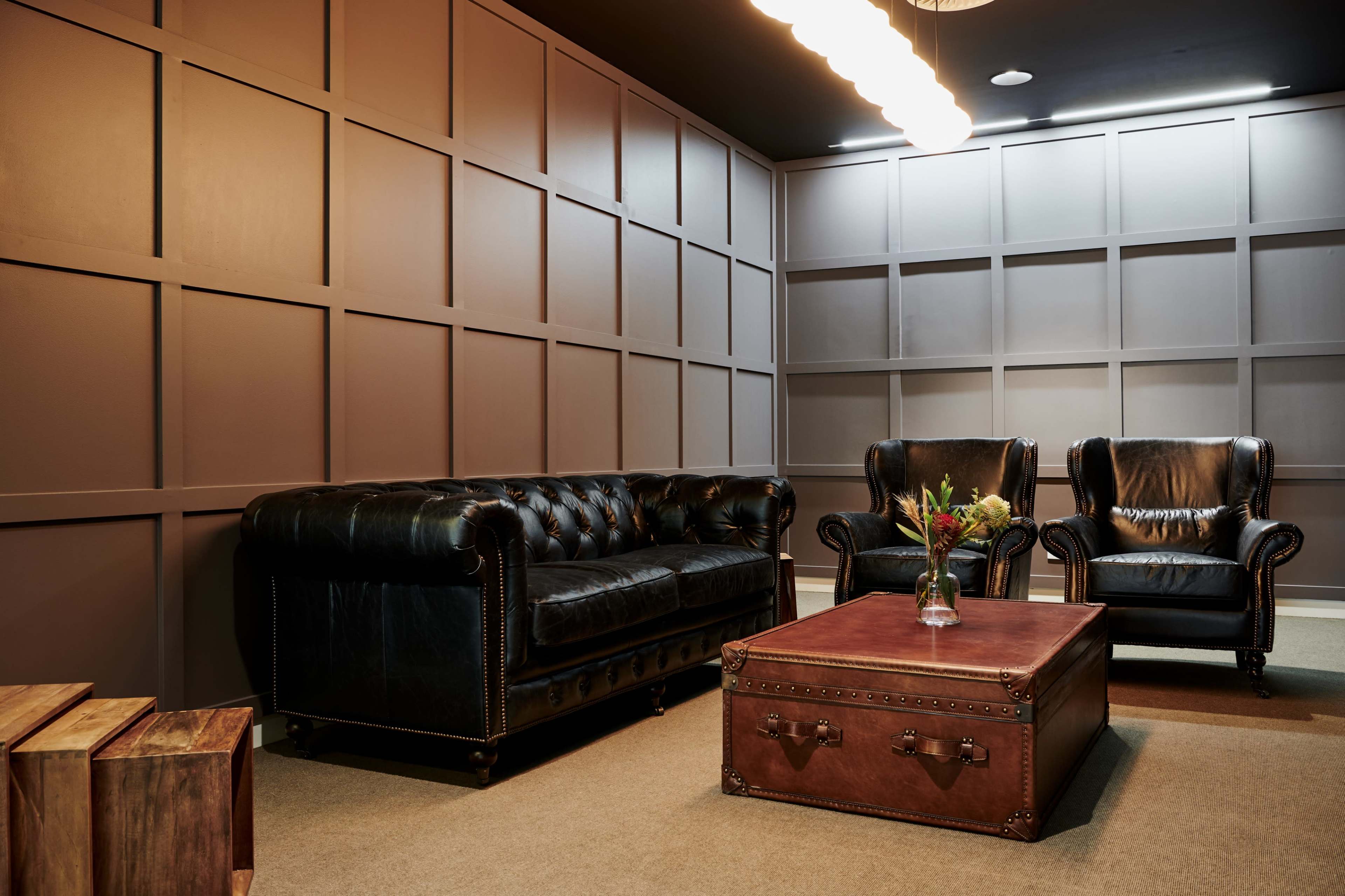 The image shows a stylish lounge area featuring a black leather chesterfield sofa, two black leather armchairs, and a brown coffee table with metal accents.