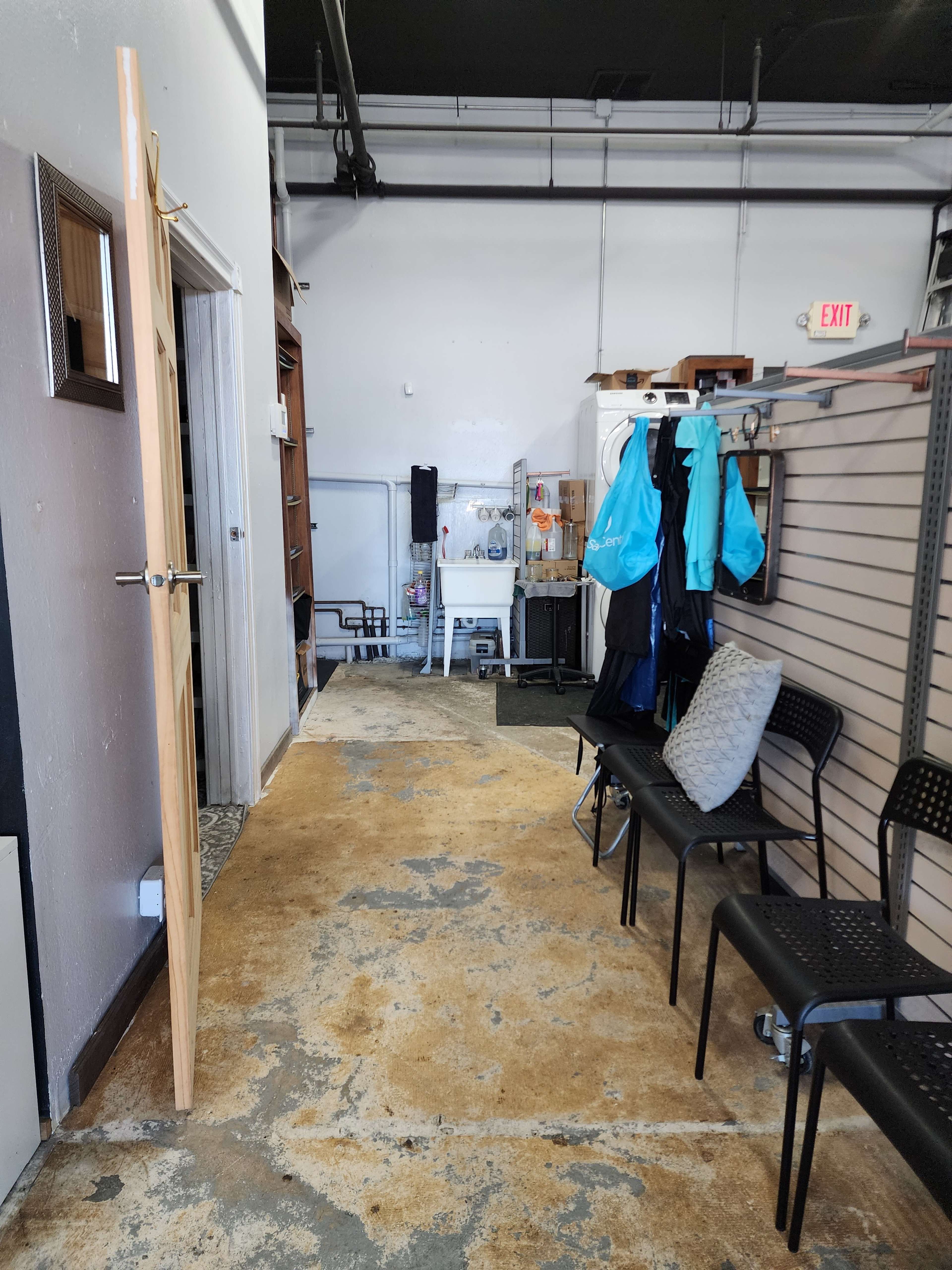 A hallway with a wooden door on the left and several black chairs lining the right side, leading to a room filled with various furniture and equipment.