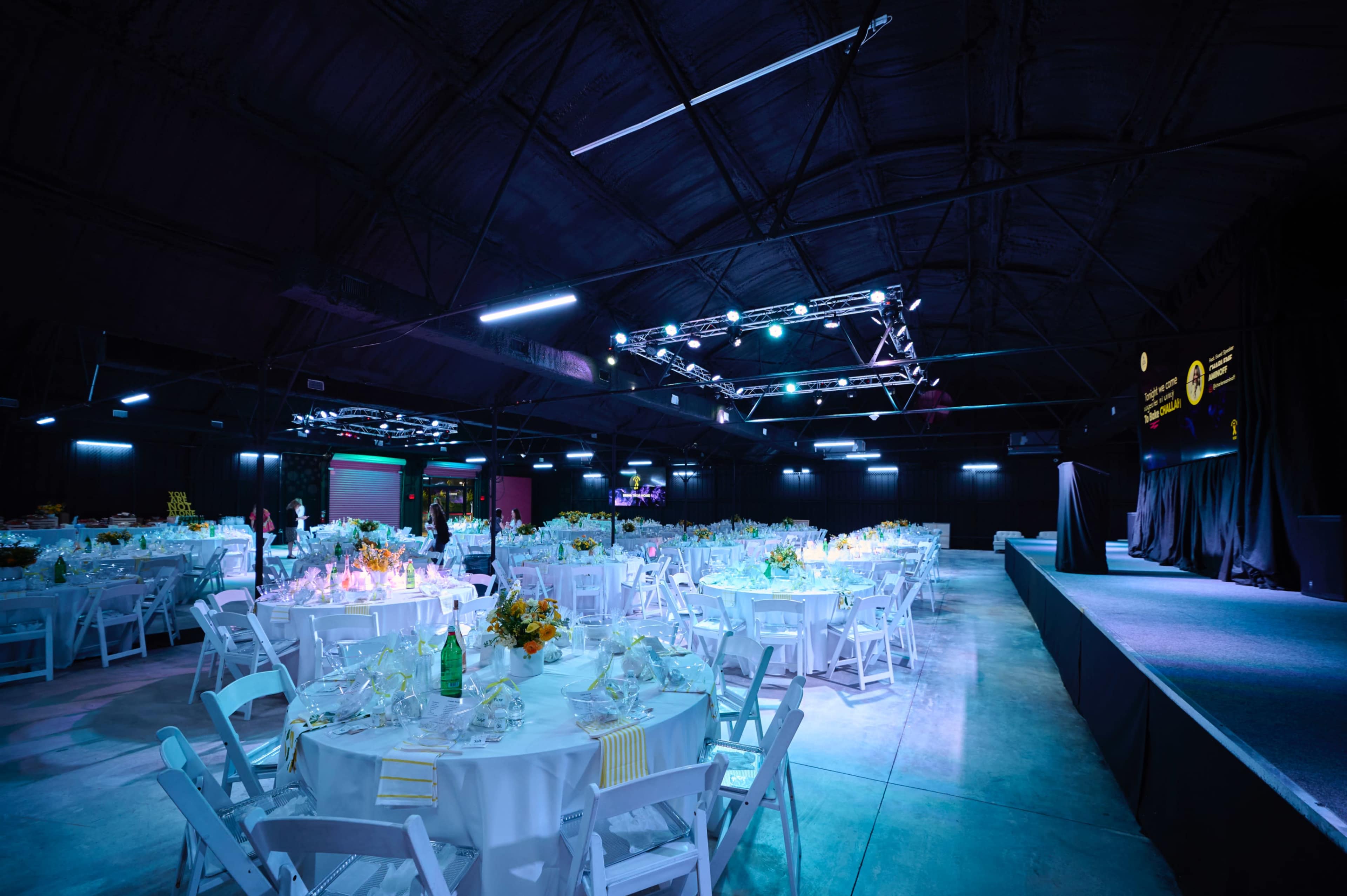A large event space is set up with multiple round tables covered in white tablecloths, each adorned with floral centerpieces and dinnerware, surrounded by soft blue lighting.