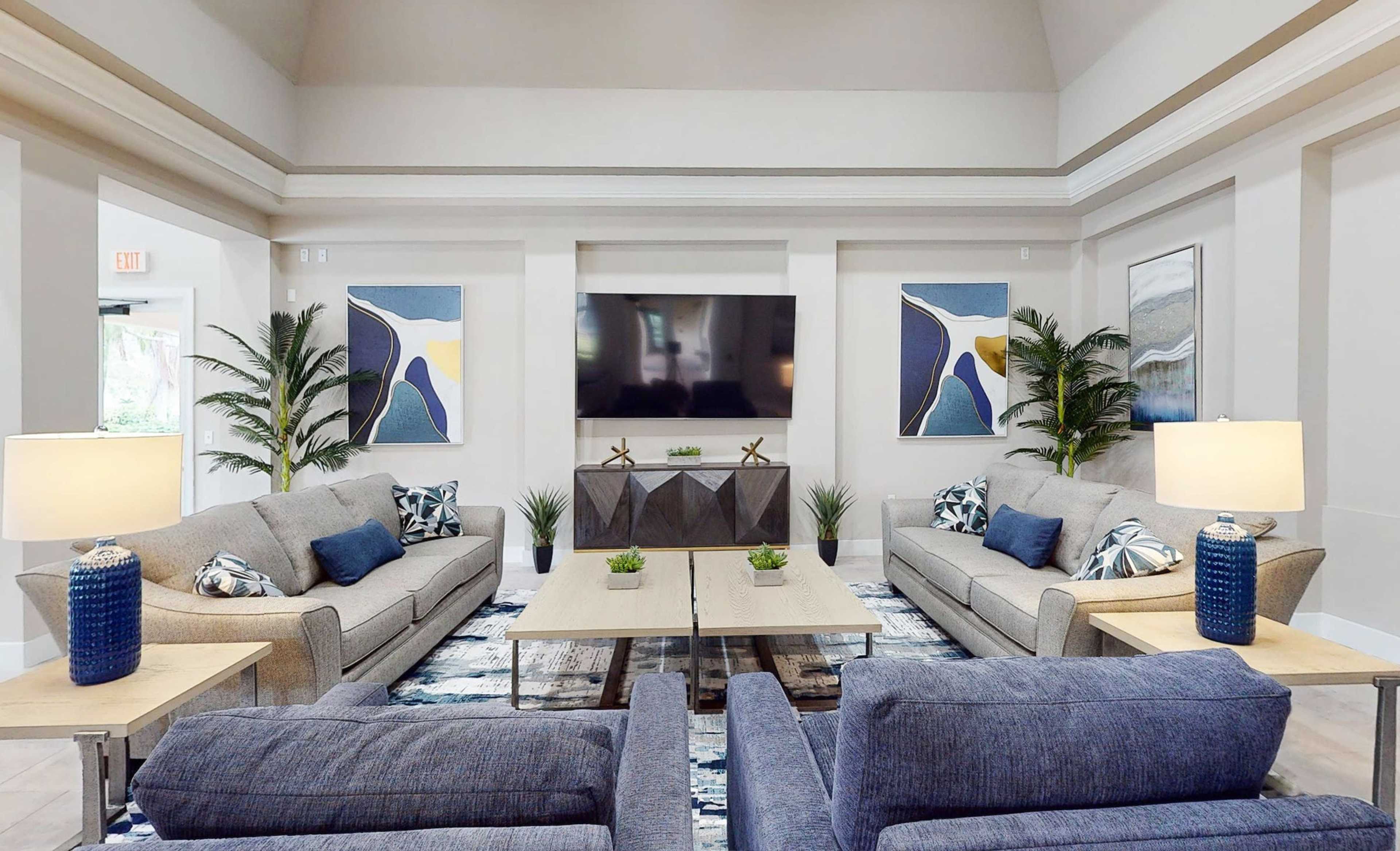 The image shows a spacious modern living area with two gray sofas and accent chairs arranged around coffee tables, featuring plants and abstract artwork on the walls.
