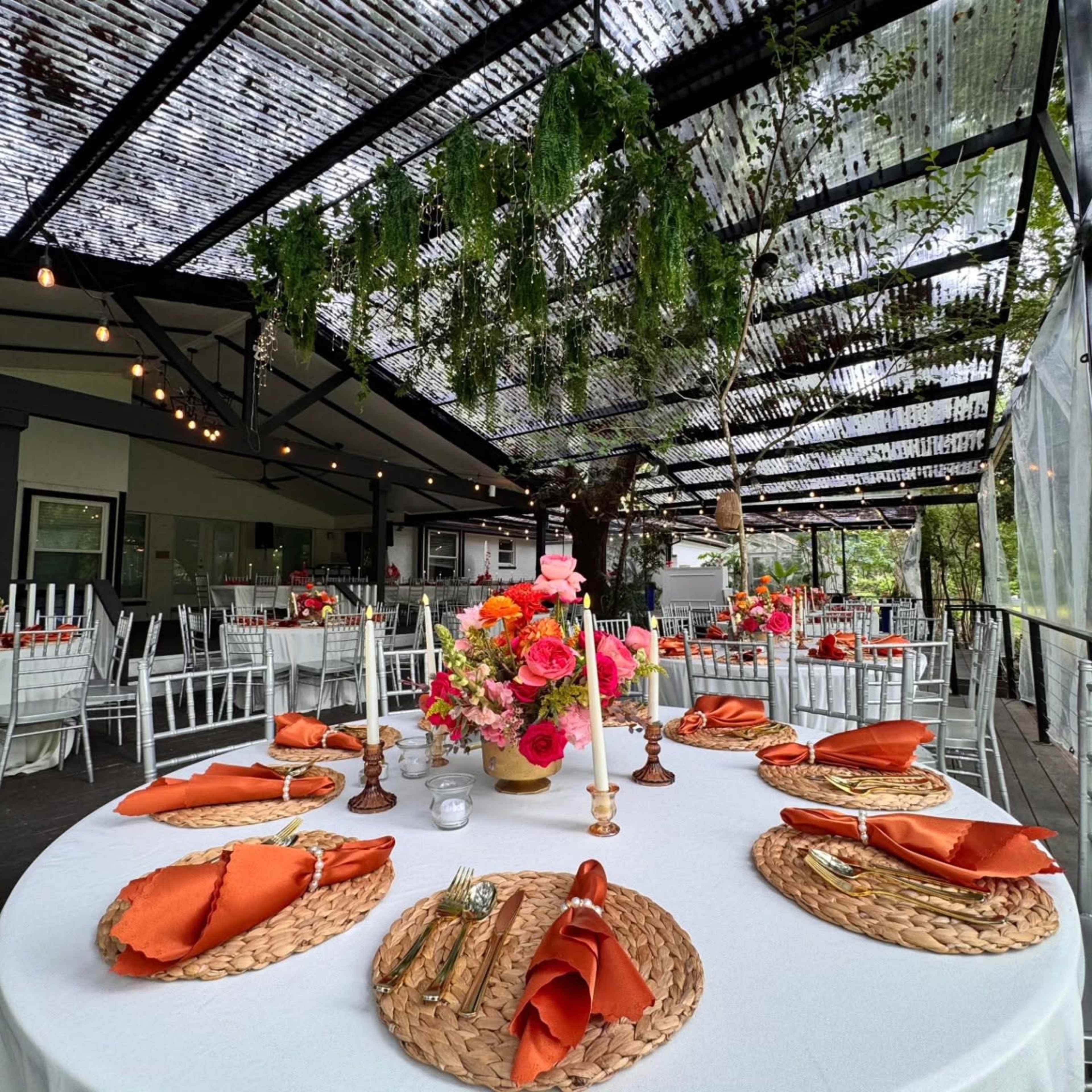 A beautifully set dining area features round tables with white tablecloths, orange napkins, floral centerpieces, and decorated outdoor seating under a canopy.