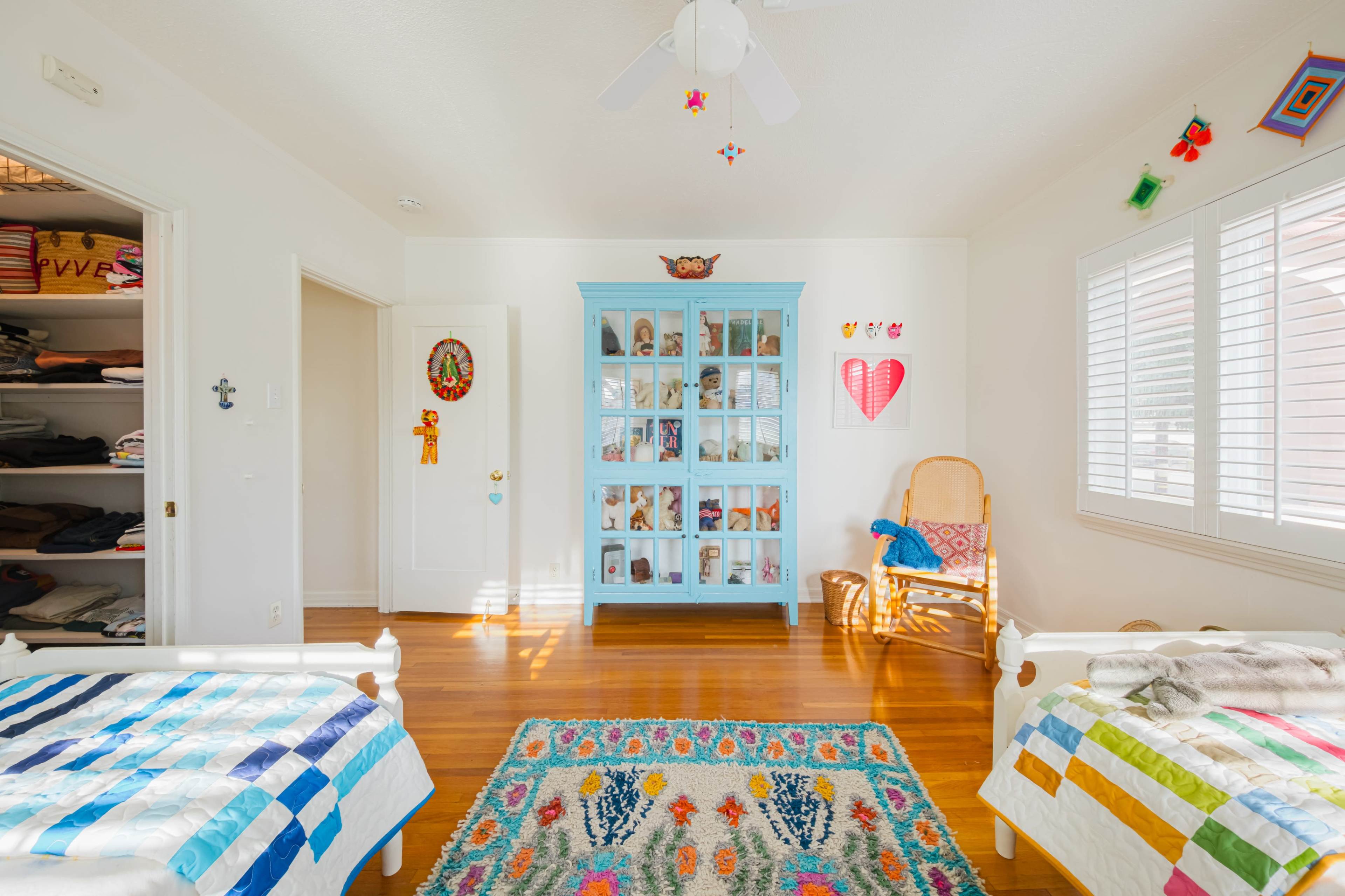 The image shows a brightly lit bedroom with two twin beds, a blue display cabinet, and a colorful area rug on a hardwood floor.