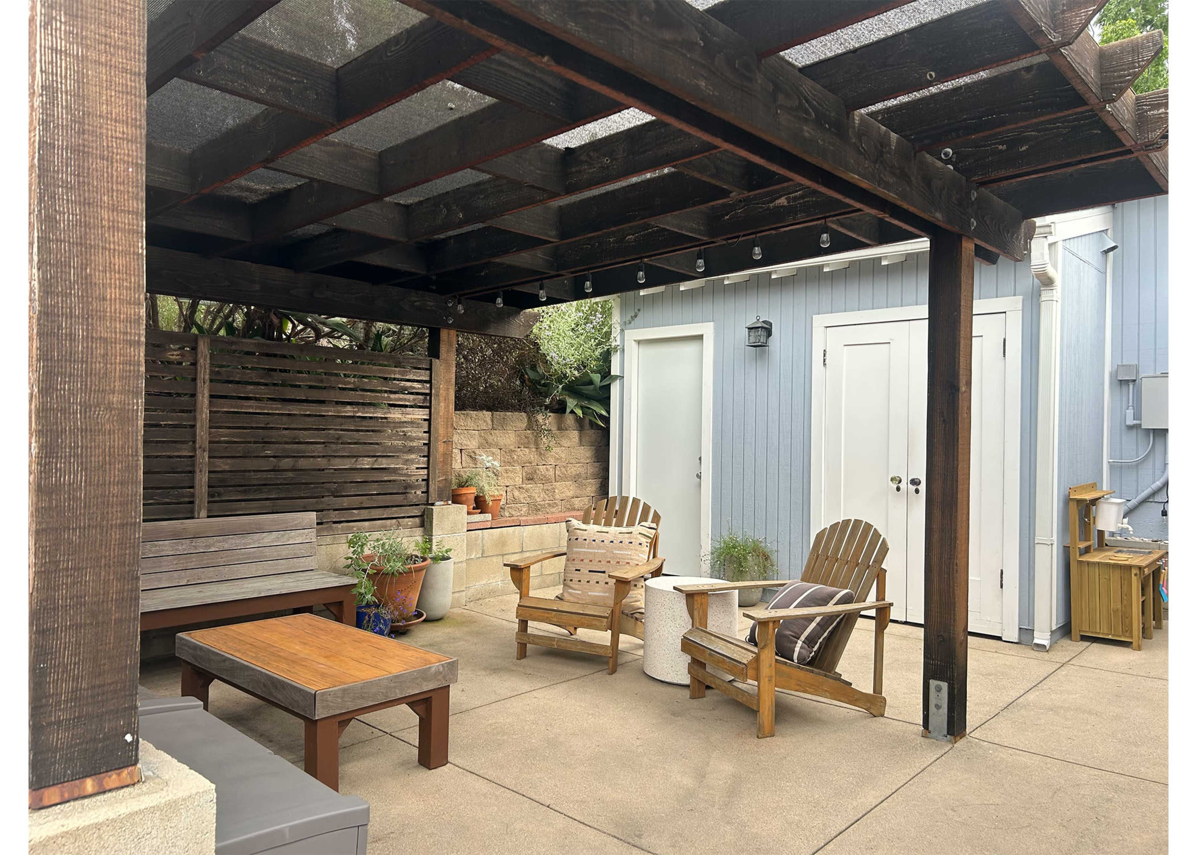 The image shows a patio area with a wooden pergola, featuring a seating arrangement that includes two wooden chairs and a table.