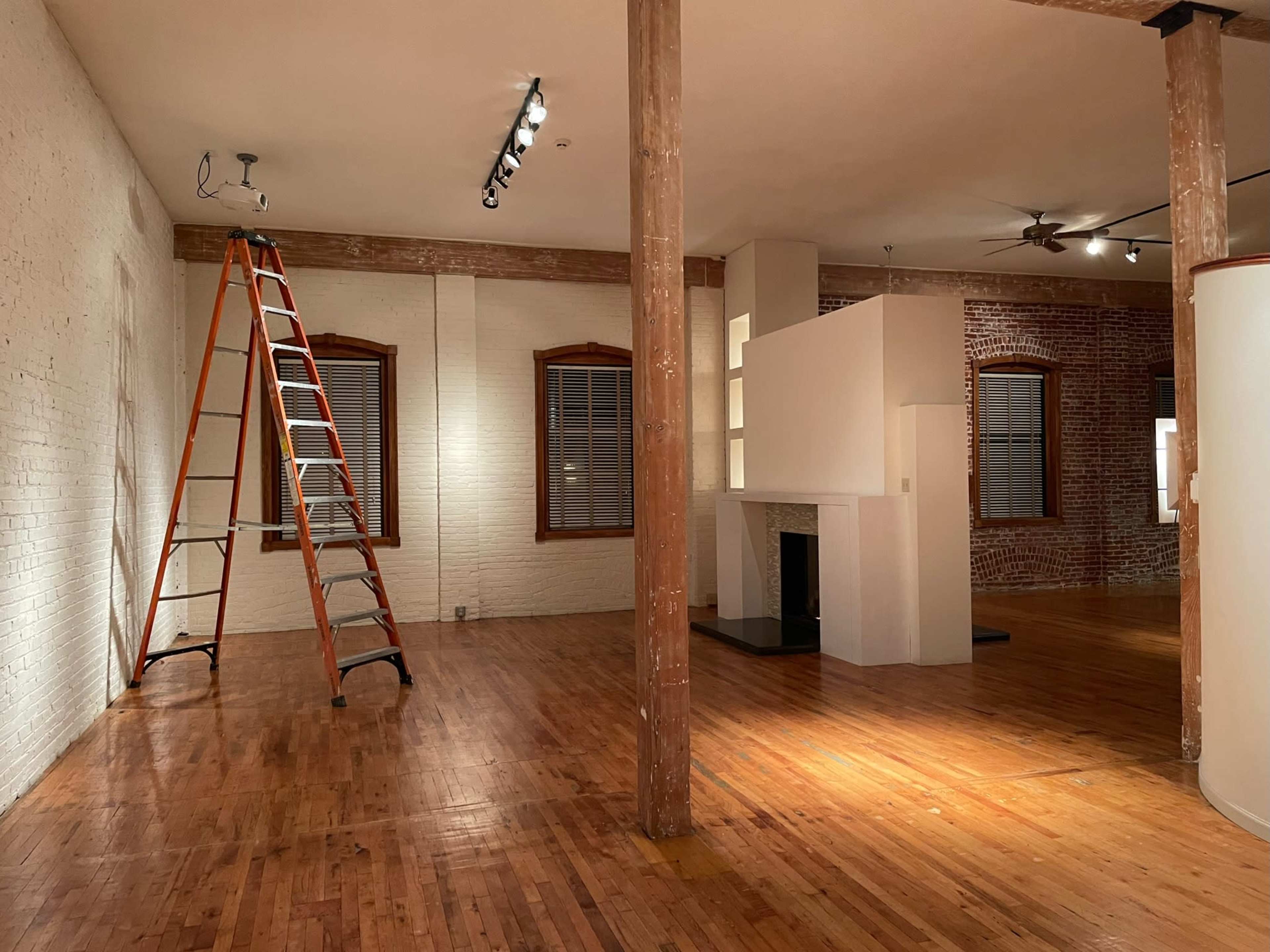 An empty room with wooden floors features exposed brick walls, a white fireplace, a ladder, and track lighting.