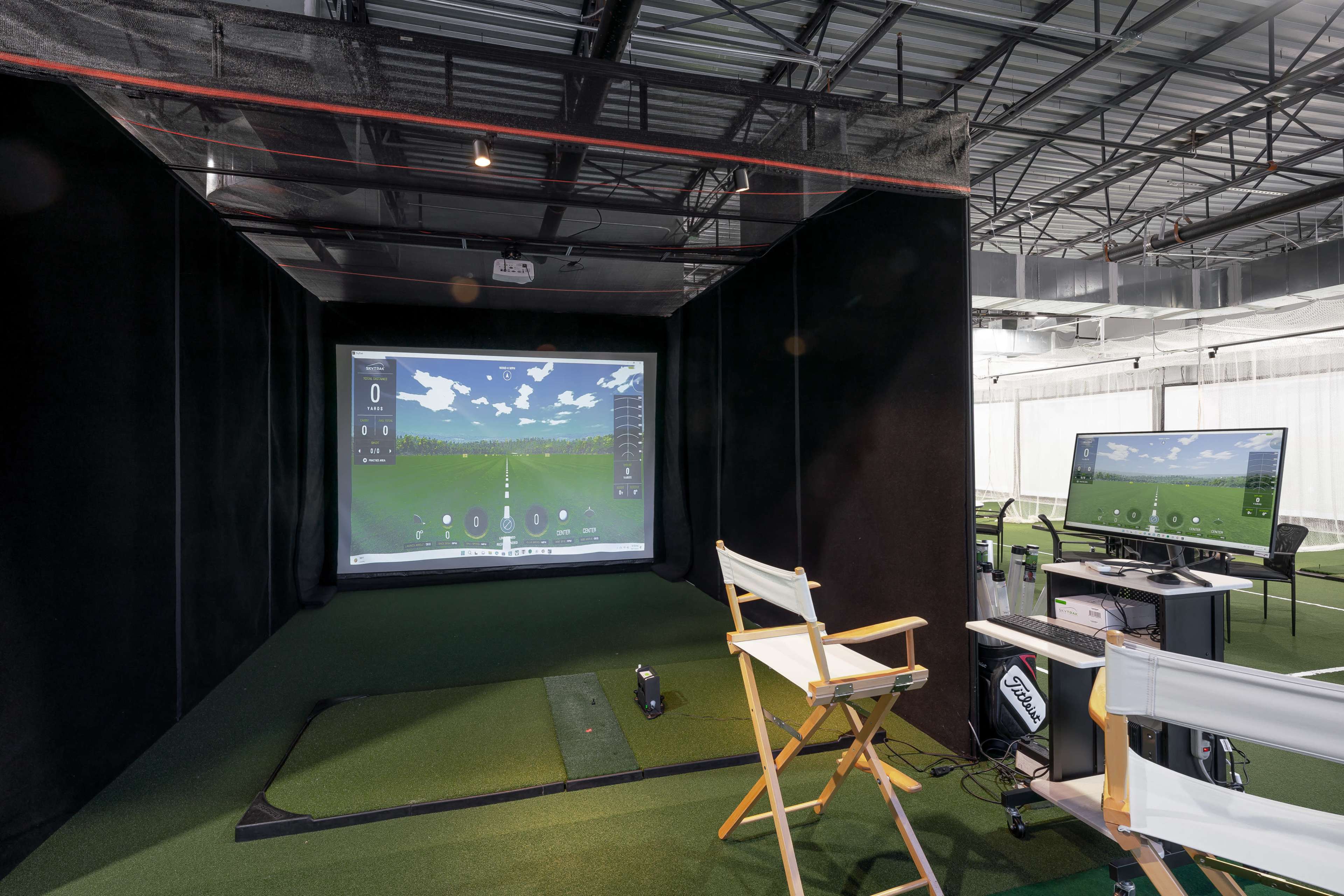 An indoor golf simulator setup with a large projection screen displaying a virtual golf course and a chair nearby.