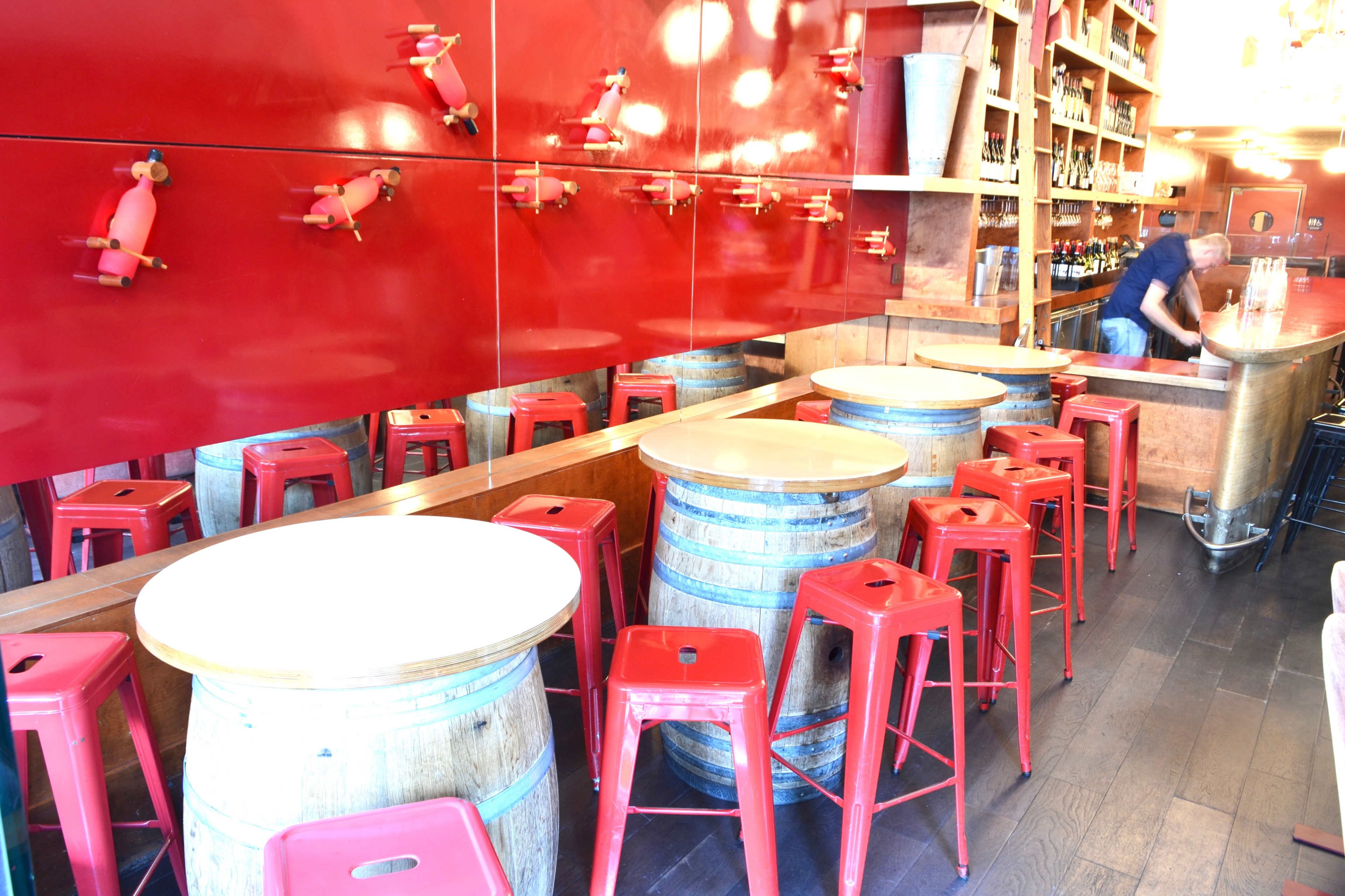 A bar area with wooden barrel tables, red metal stools, and a bright red wall behind the bar shelves filled with bottles.