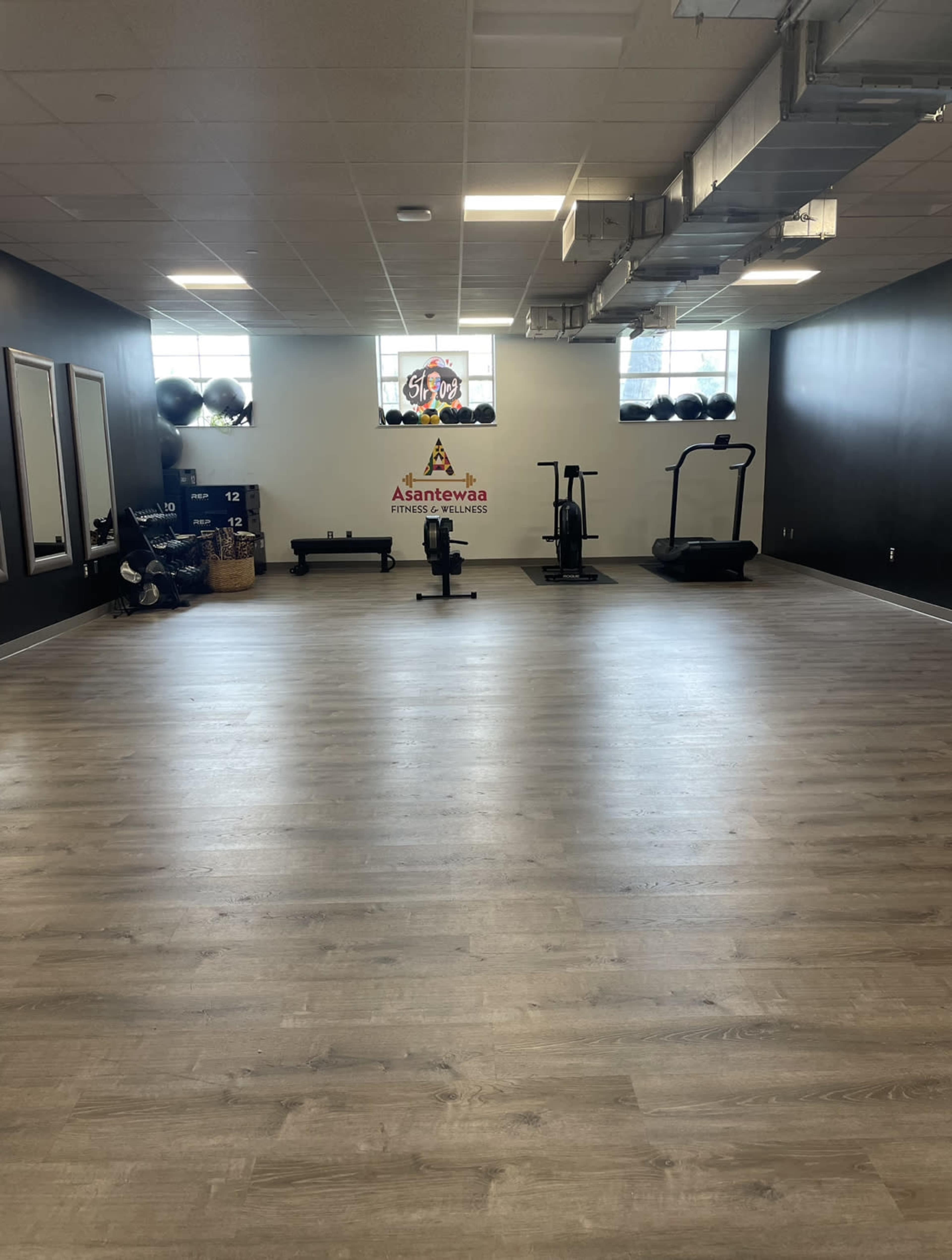 A spacious gym with a wooden floor, exercise equipment like a treadmill and weights, and large windows letting in light.