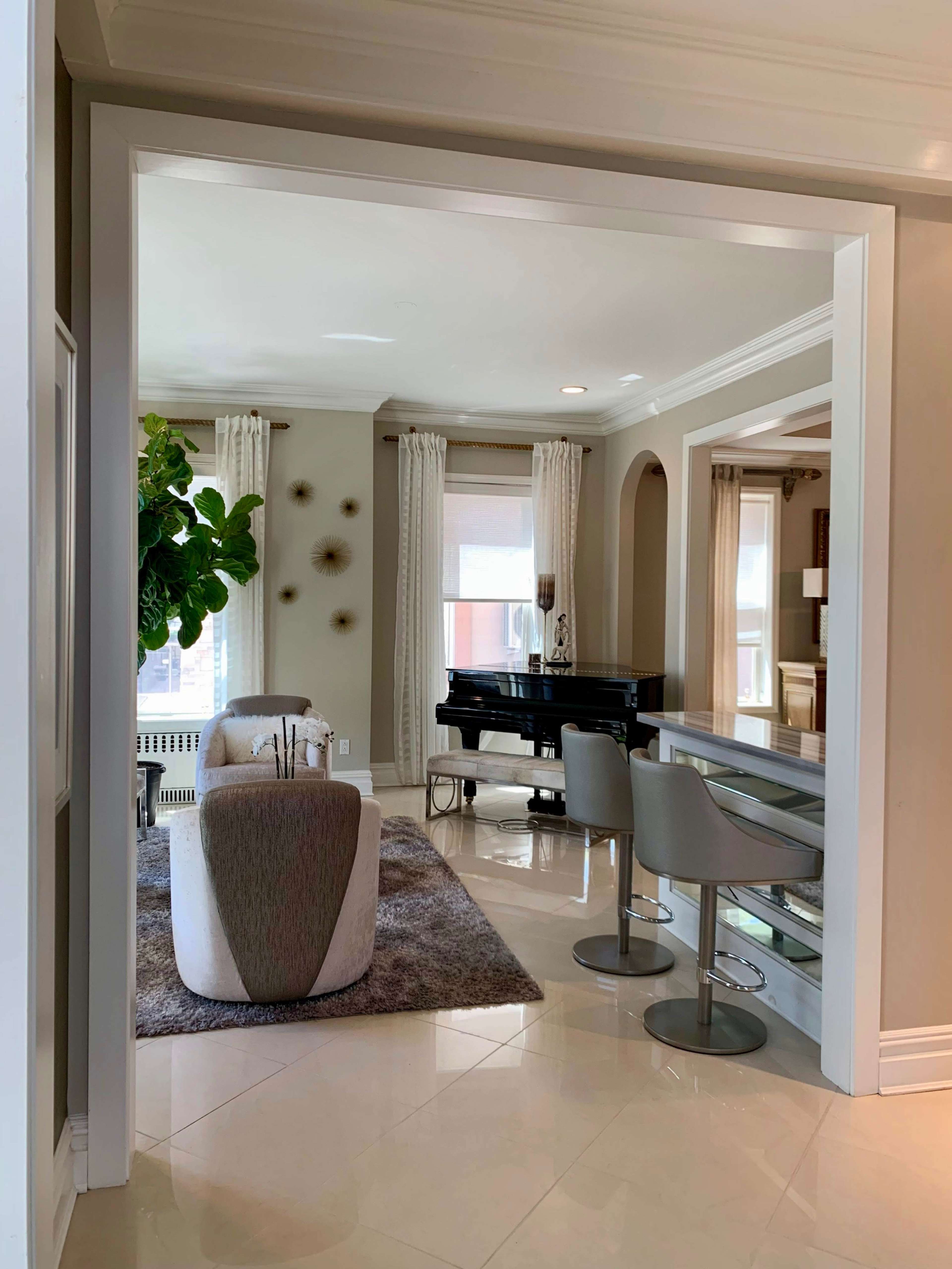 A modern living space featuring a grand piano, dining area with barstools, and large windows framed by curtains.