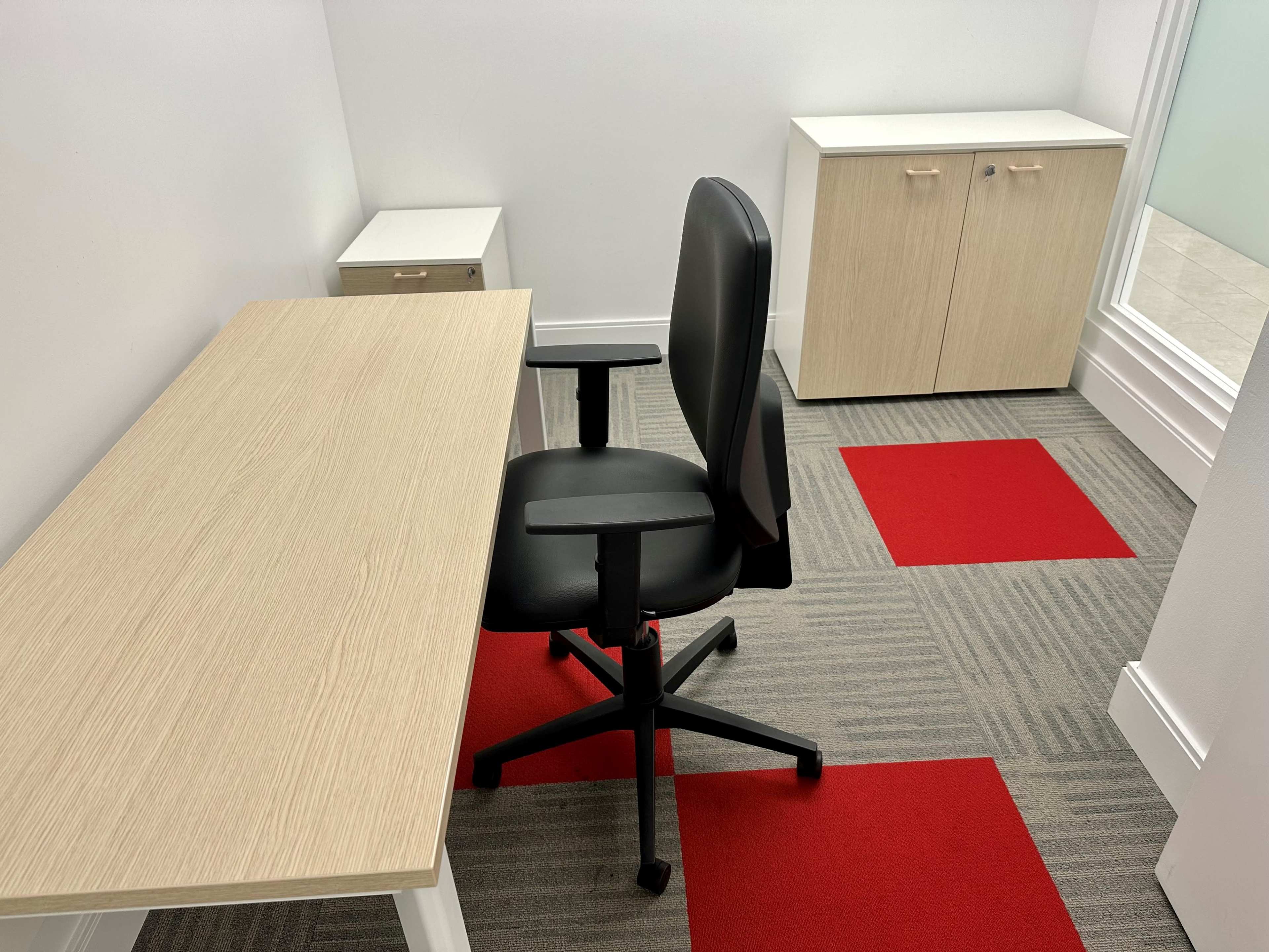 A small office space featuring a light wood desk, a black office chair, and two cabinets, with red carpet tiles on the floor.