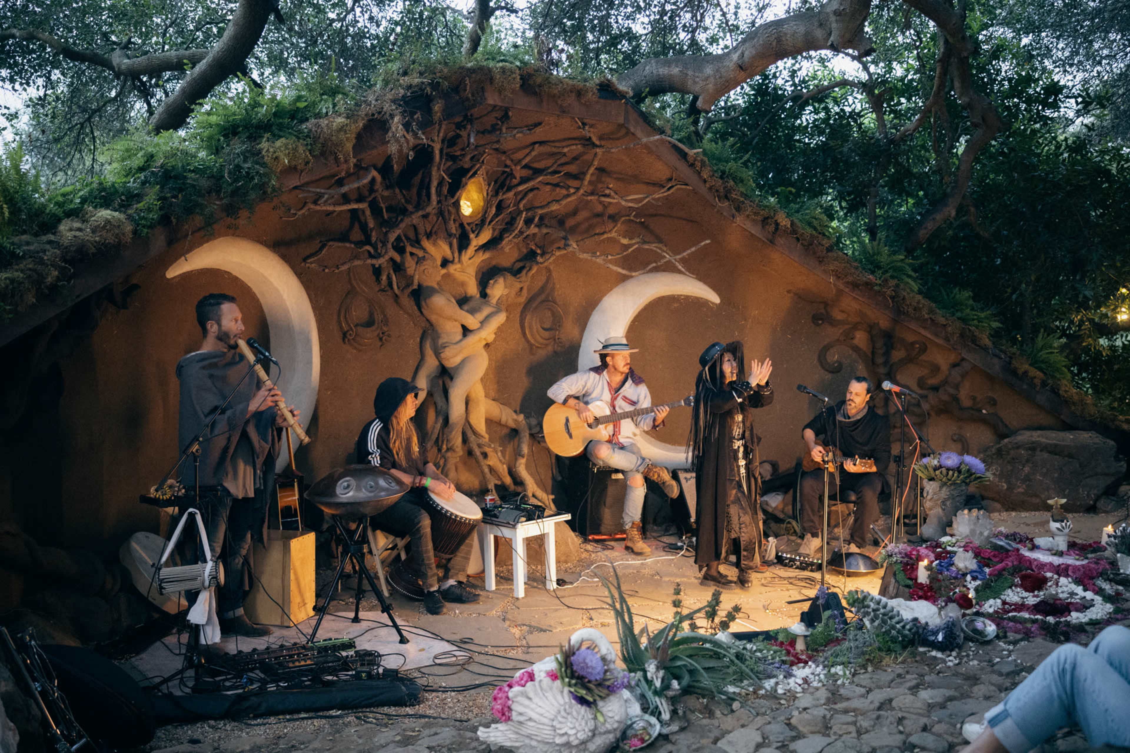 A group of five musicians performs together in a natural setting, surrounded by decorative foliage and a rustic backdrop featuring moon motifs.