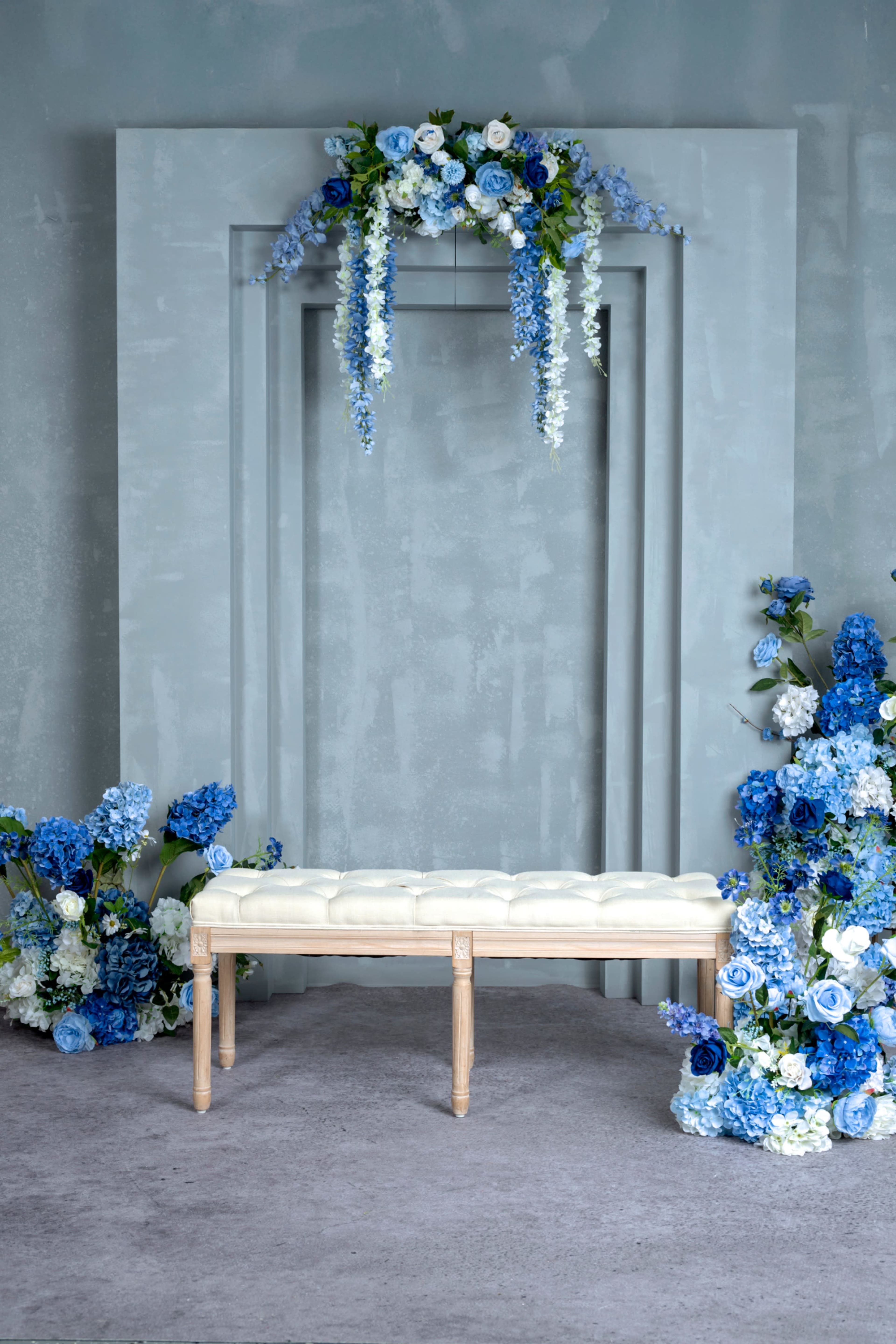 The image features a decorative setting with a light-colored bench in front of a blue floral arrangement and a backdrop of stylized geometric shapes.