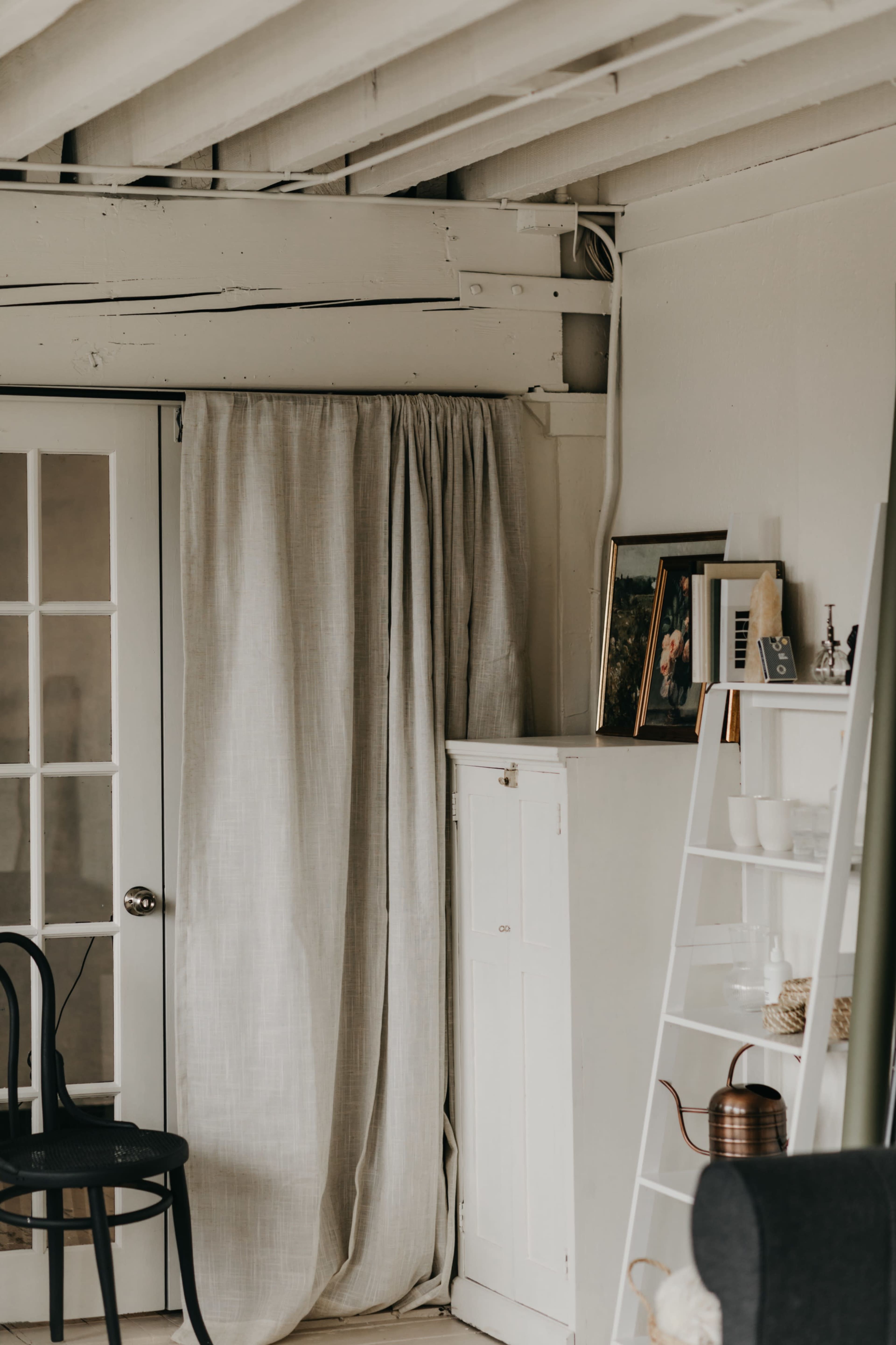 The image shows a corner of a room featuring a white curtain, a closed glass-paned door, a white storage cabinet, and a leaning shelf with decorative items.
