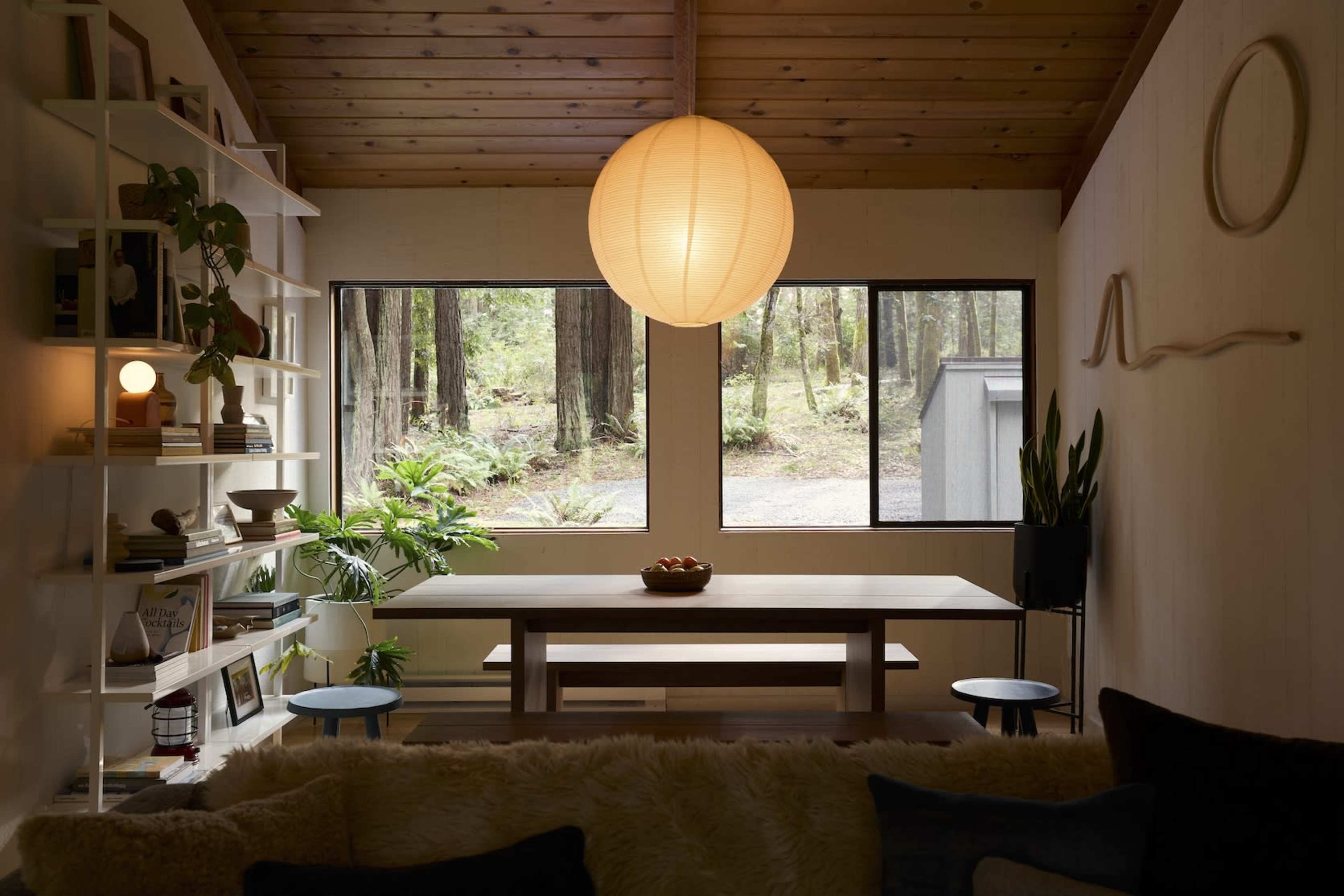 A dining area features a wooden table surrounded by stools, illuminated by a large pendant light, with windows showcasing a forest view.