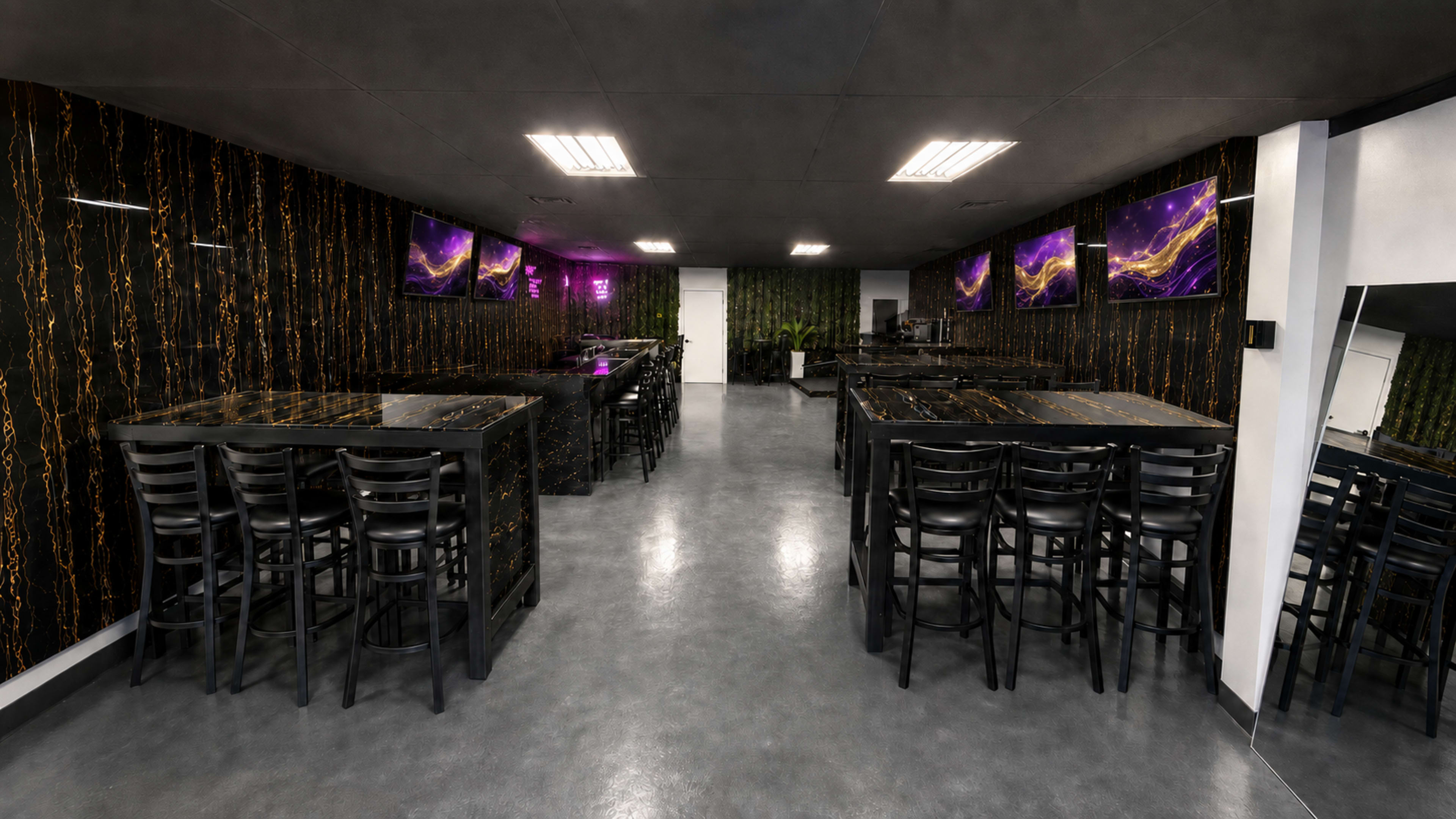 The image shows a spacious, dimly lit interior with black tables and chairs arranged in rows, accented by textured walls and multiple screens displaying vibrant visuals.