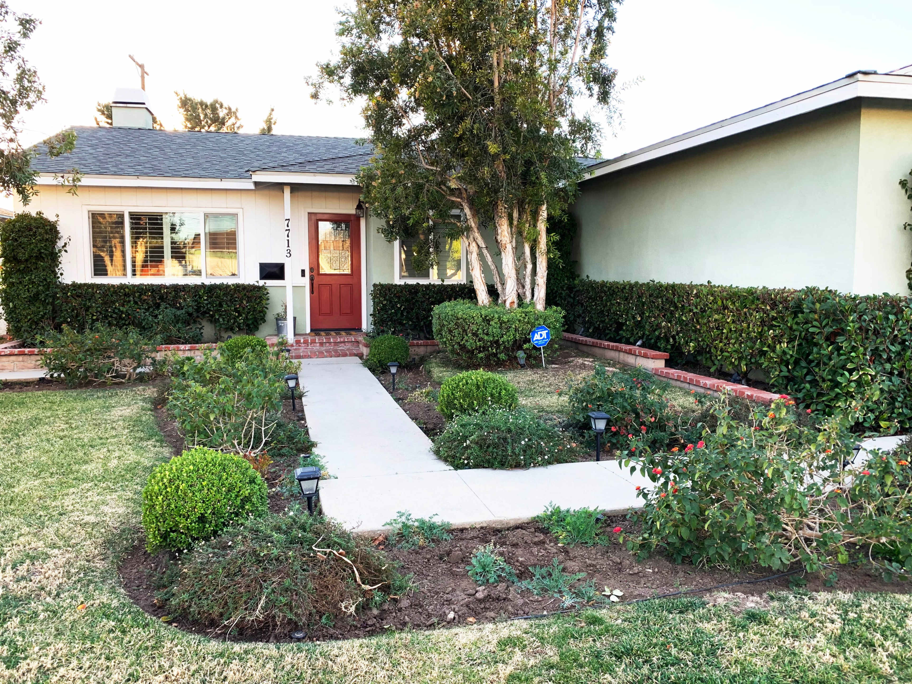A well-maintained front yard features a concrete pathway leading to a house with a red door, surrounded by neatly trimmed shrubs and garden beds.
