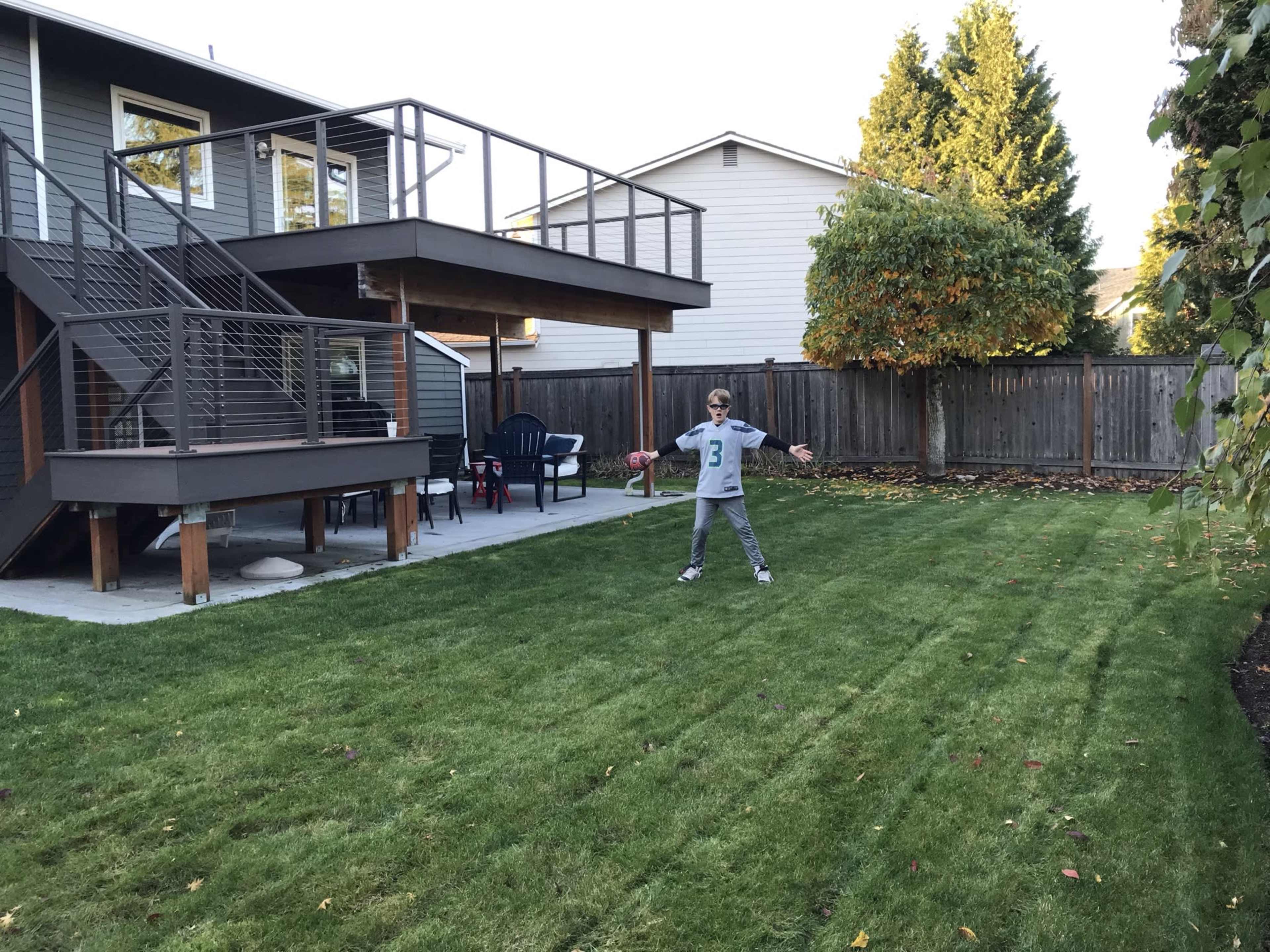 A child stands in a neatly mowed backyard with a deck in the background and a few lawn chairs nearby.