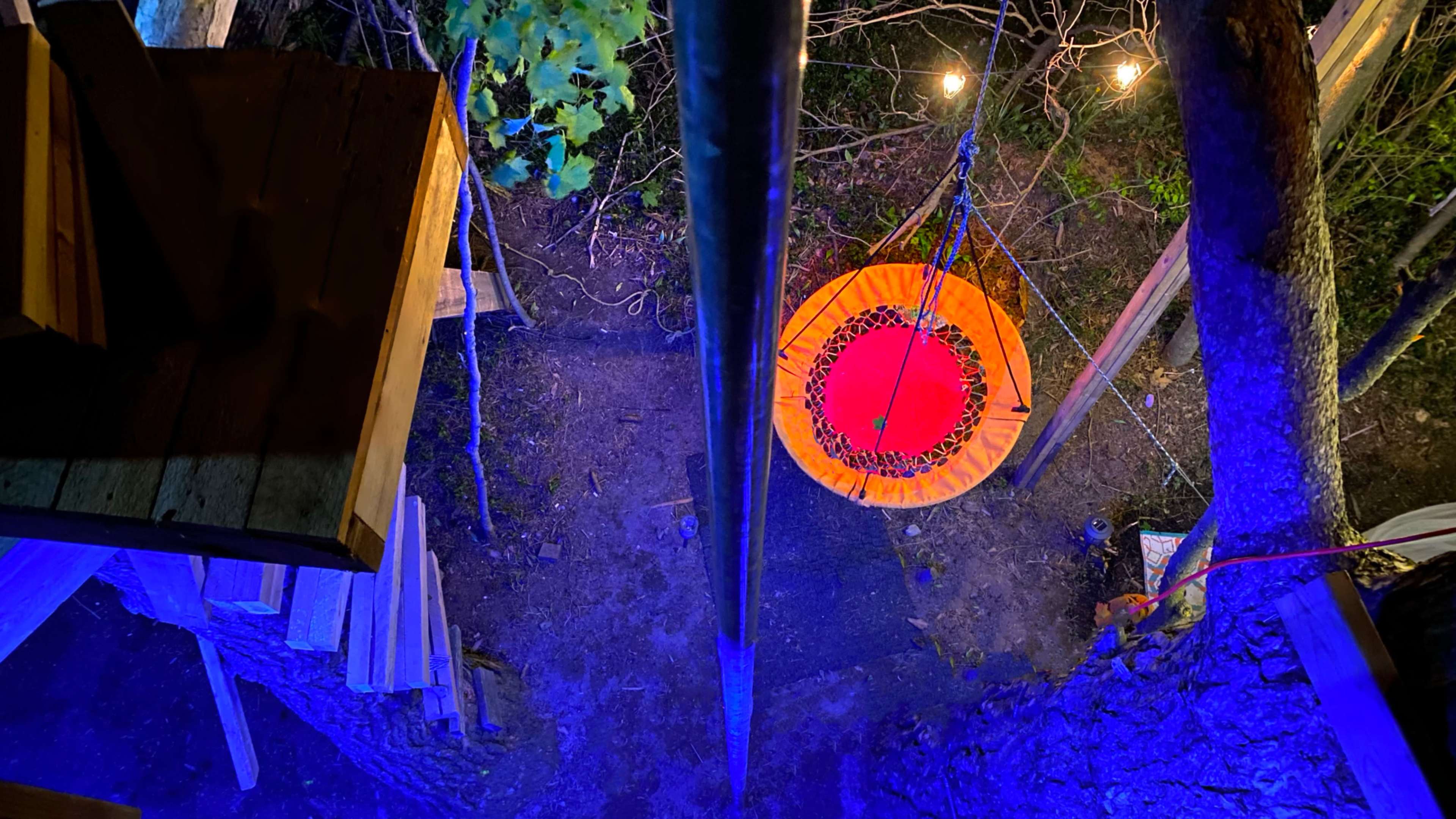 The image shows a colorful, round swing hanging from a tree, viewed from above, with wooden structures and lighting in the background.