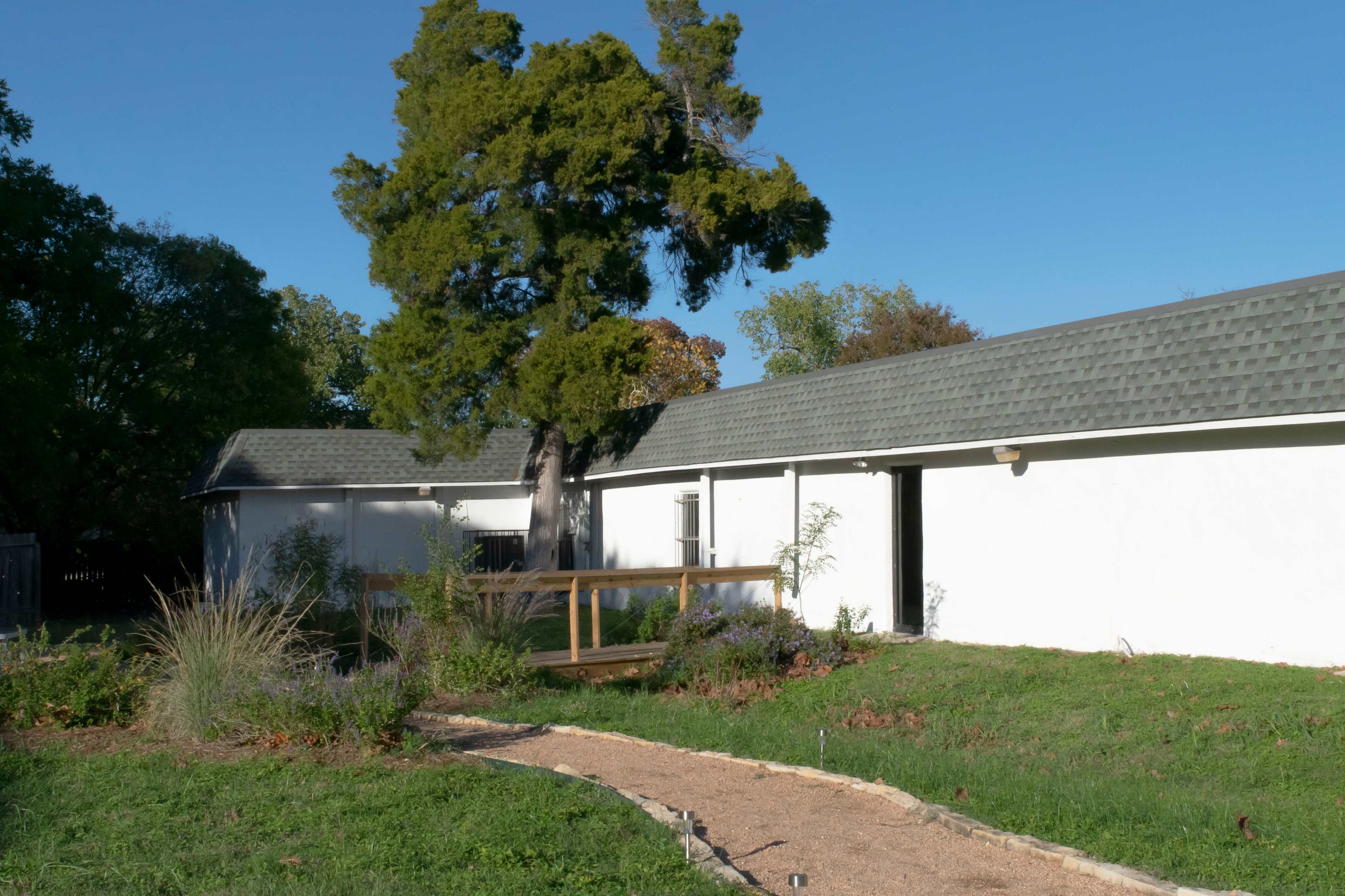 A path leads through a grassy area to a white building with a gray roof, alongside a large tree.