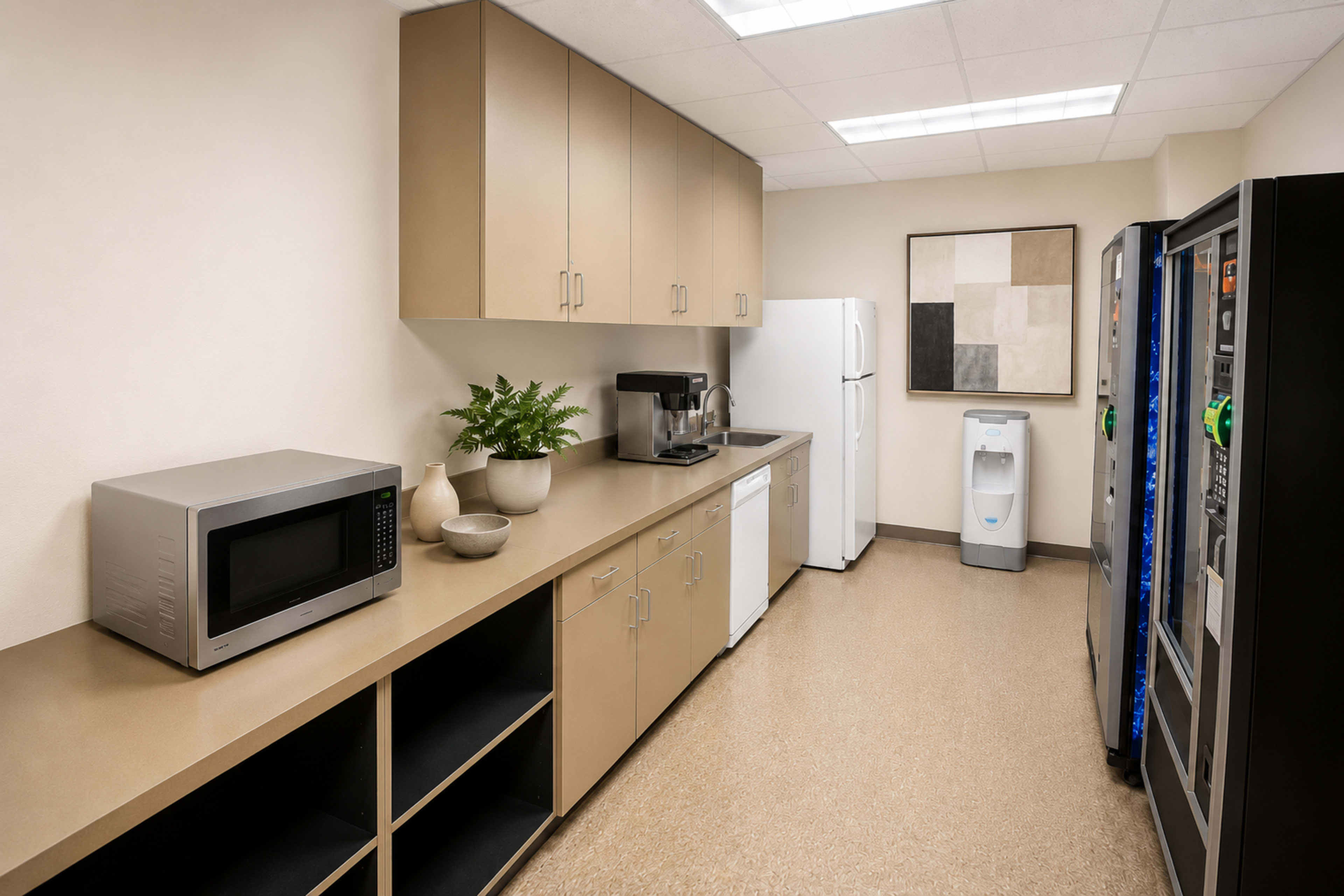 The image shows a well-organized kitchen space featuring a microwave, coffee maker, refrigerator, and vending machines along a counter with cabinets above.