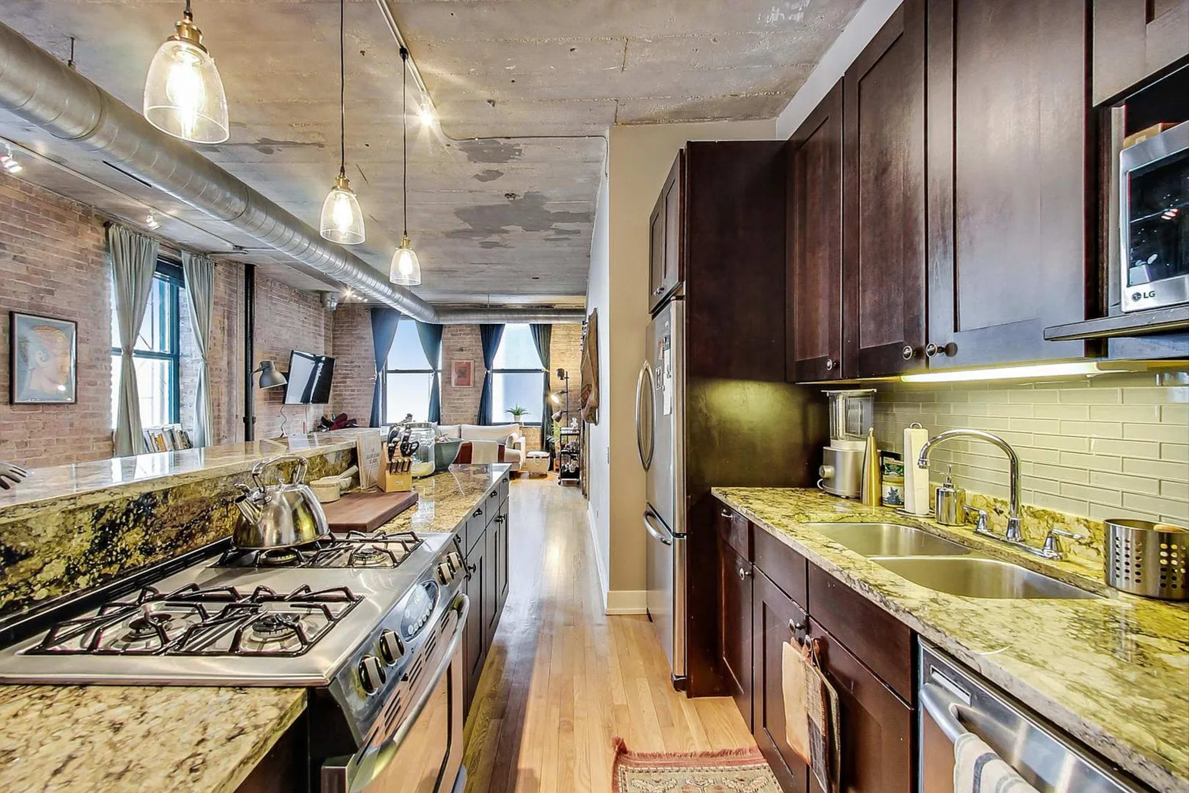The image shows a modern kitchen with dark cabinetry, a granite countertop, and stainless steel appliances, leading into a spacious living area with large windows.