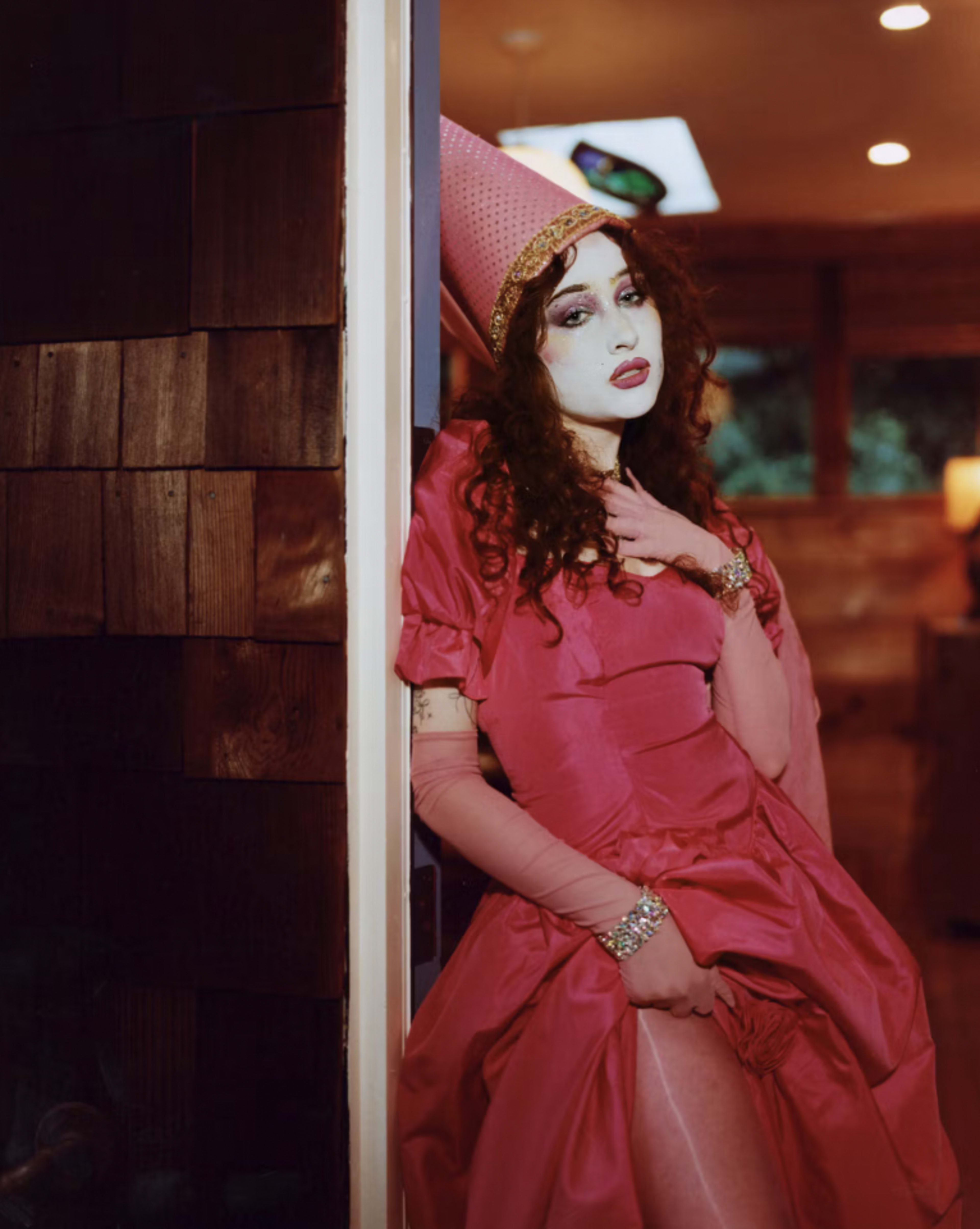 A person in a pink dress and a large pink hat stands by a doorway, posing dramatically in a modern interior space.