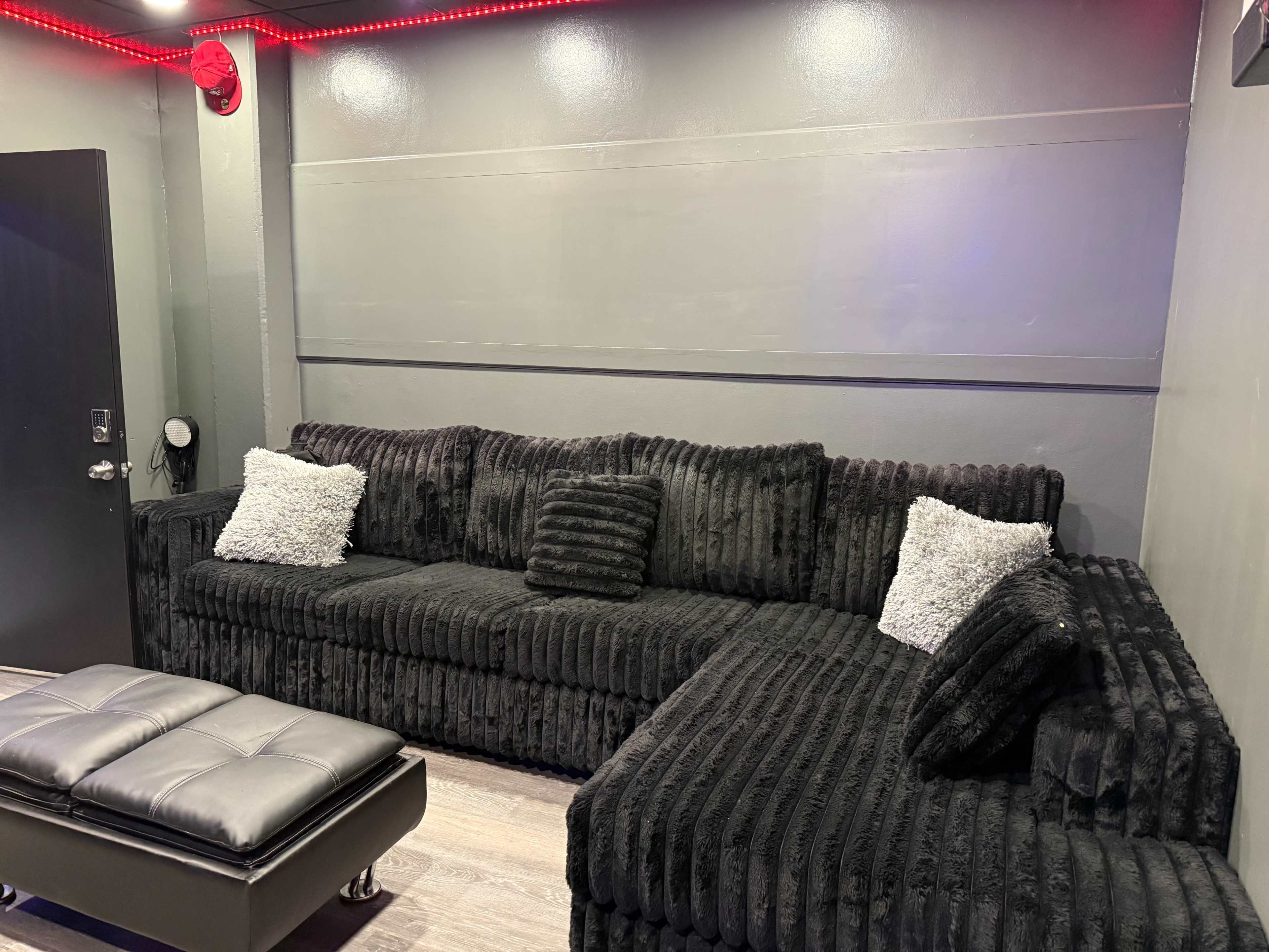 The image shows a plush, black sectional sofa with several decorative pillows against a wall in a dimly lit room.