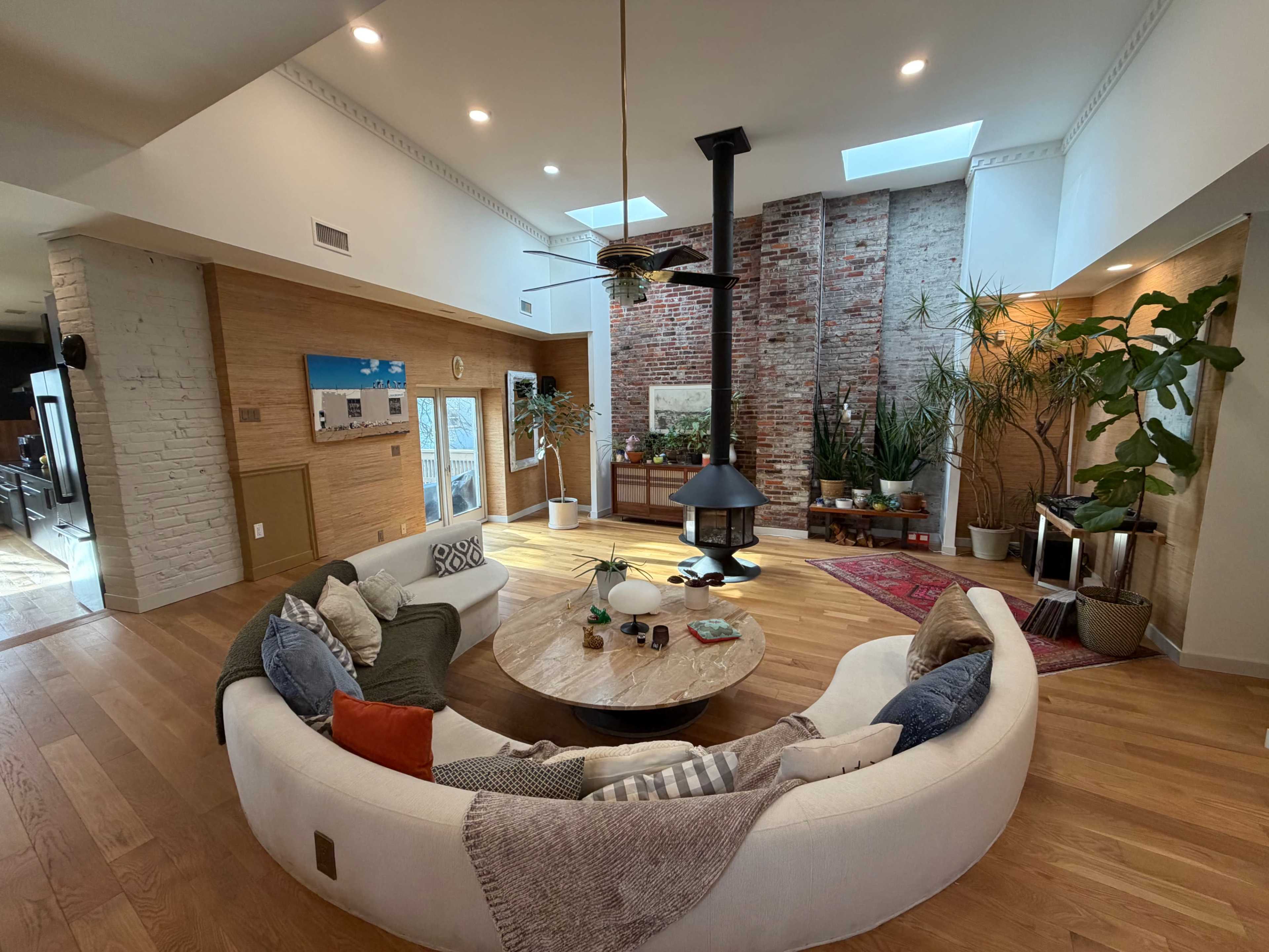 The image shows a spacious living room featuring a circular white sofa around a wooden coffee table, with a modern fireplace and large windows allowing natural light to illuminate the space.