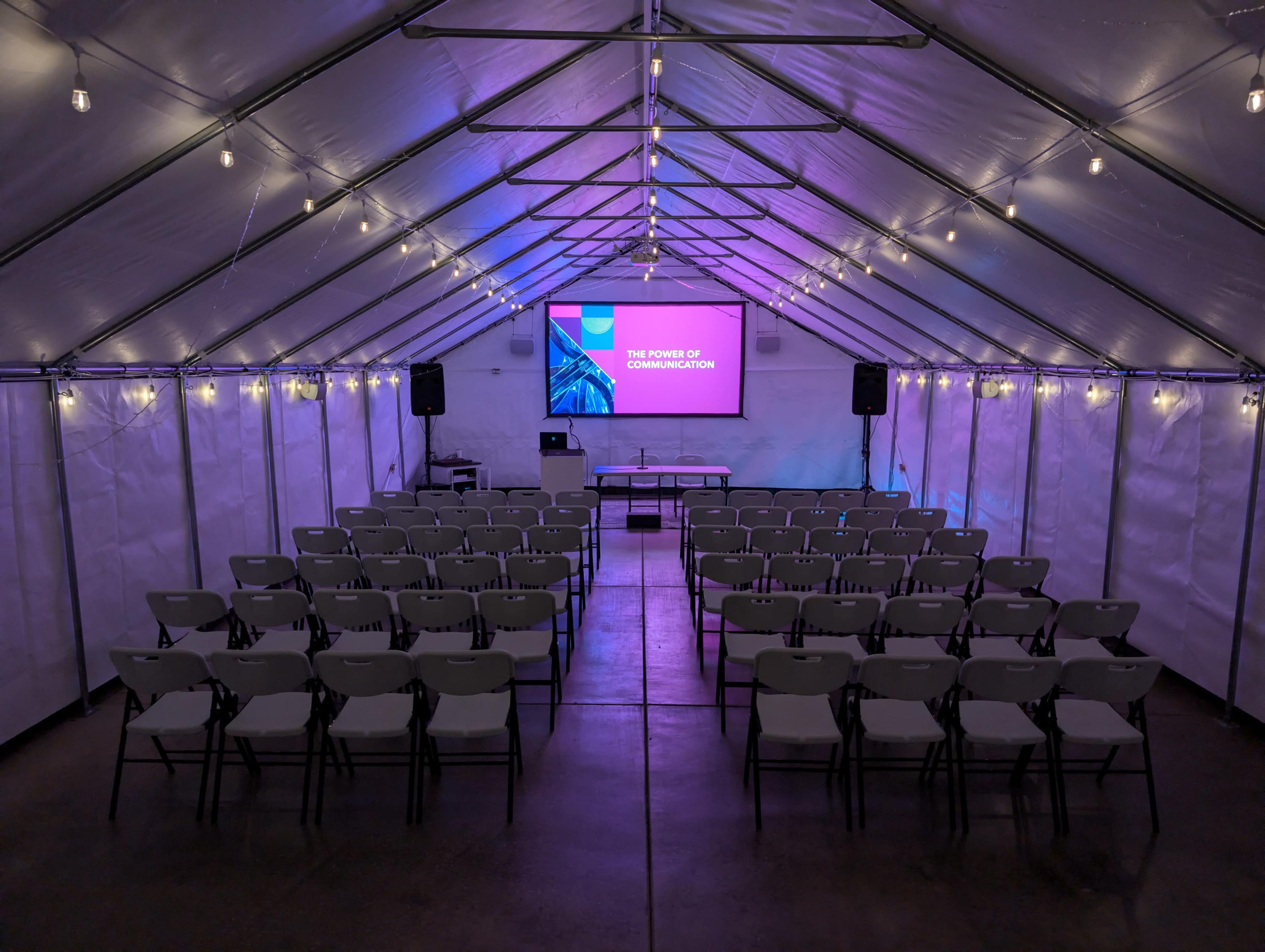 A rectangular tent is set up with rows of chairs facing a front stage displaying a presentation titled "The Power of Communication."