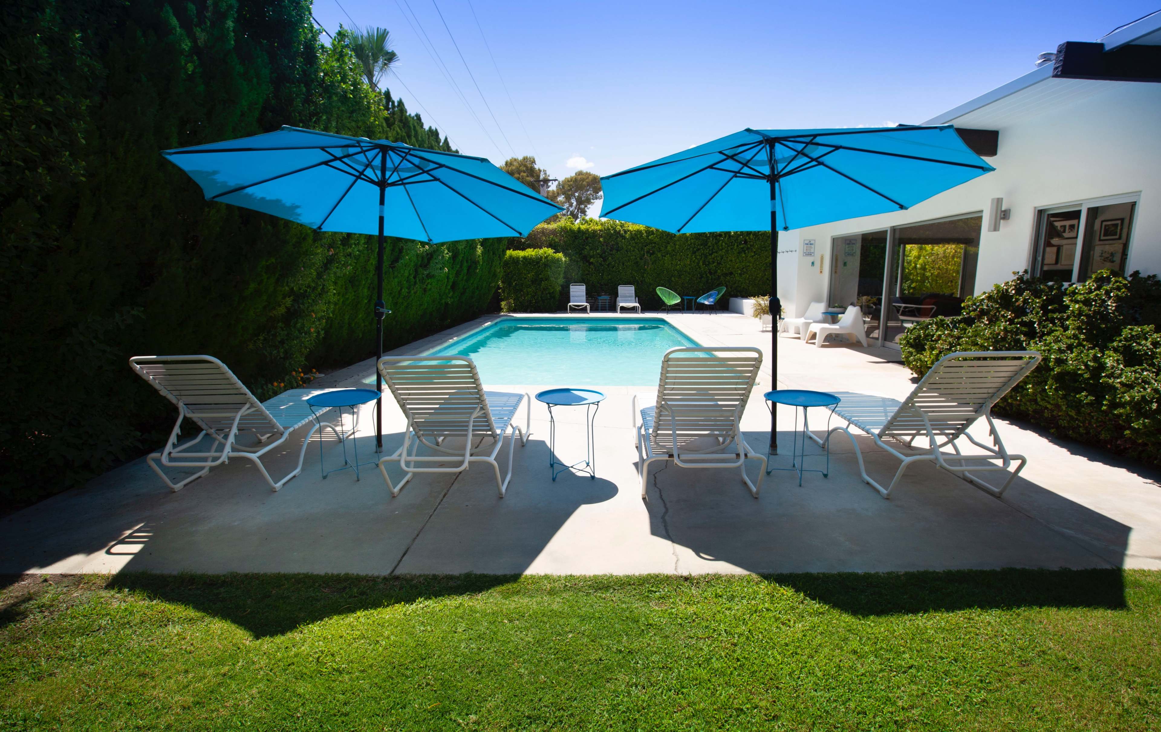 The image shows a swimming pool surrounded by lounge chairs and blue umbrellas in a landscaped backyard.