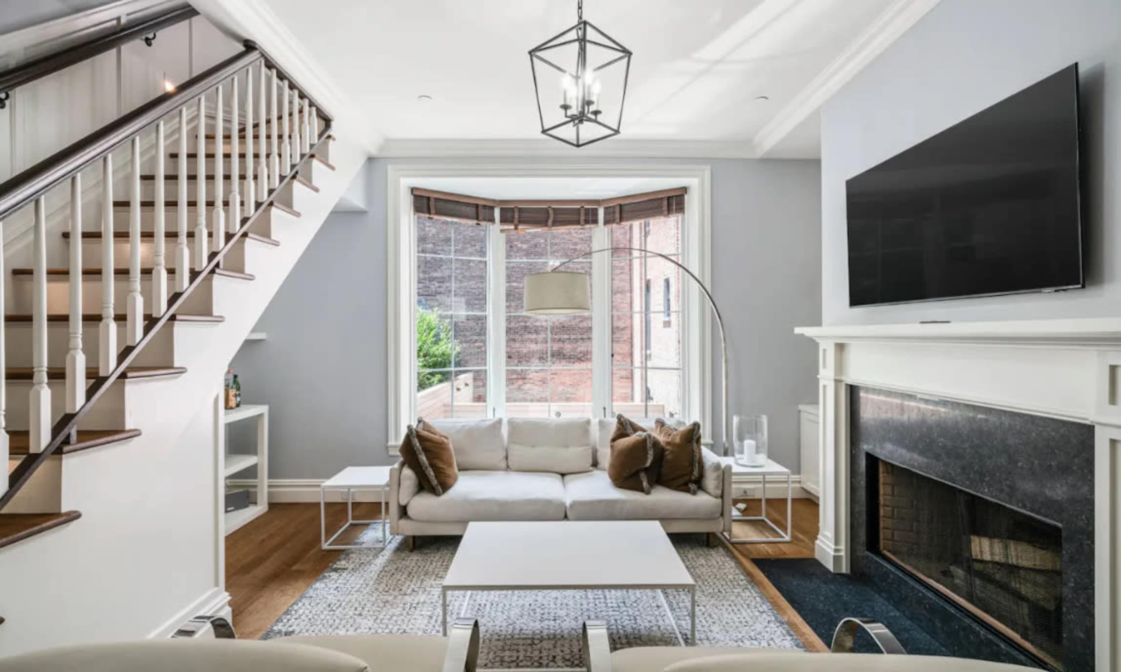 The image shows a modern living room with a staircase, large windows, a sectional sofa, and a sleek fireplace.