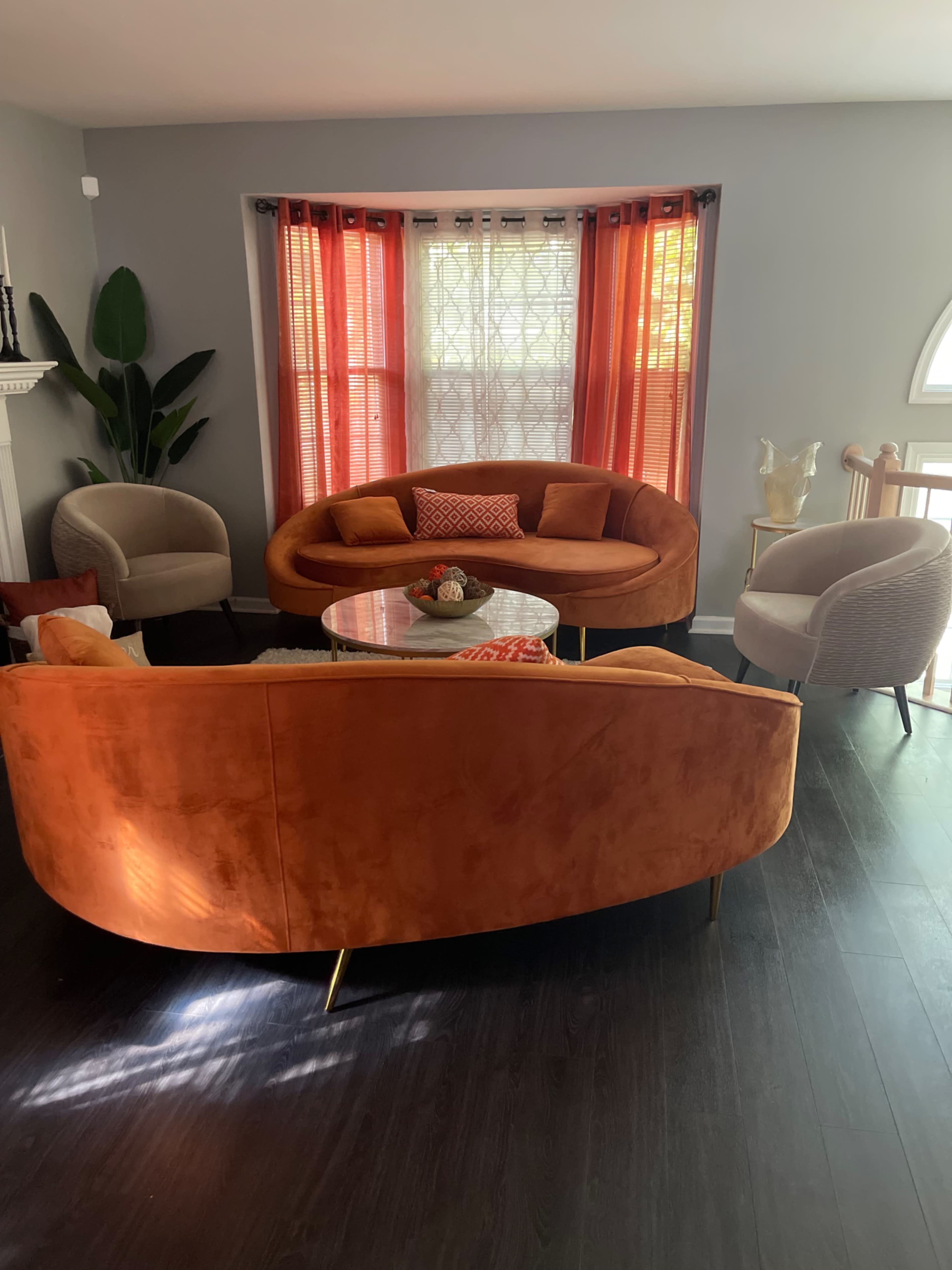 A brightly lit living room features an orange velvet curved sofa, a round glass coffee table, and a mix of accent chairs, with large windows draped in red curtains.