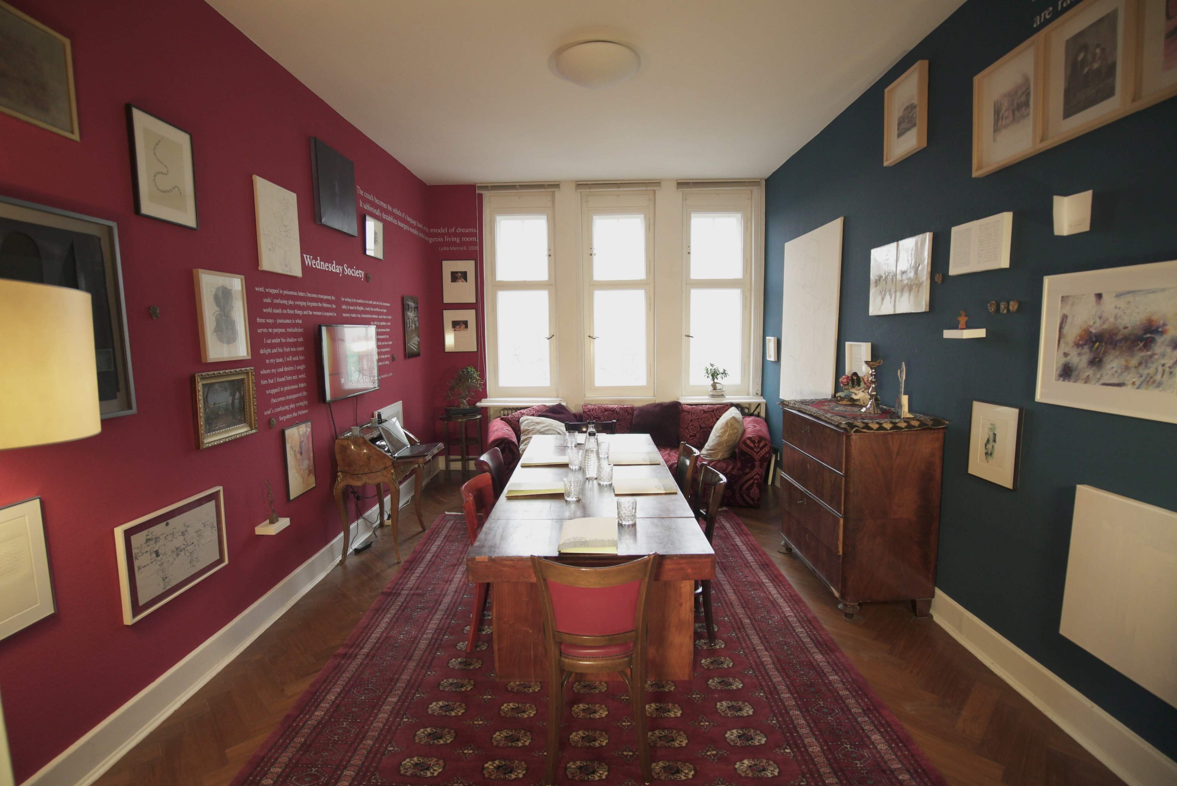 The image shows a well-decorated dining room with a long wooden table surrounded by chairs, against a backdrop of red and blue walls adorned with framed artwork and photographs.