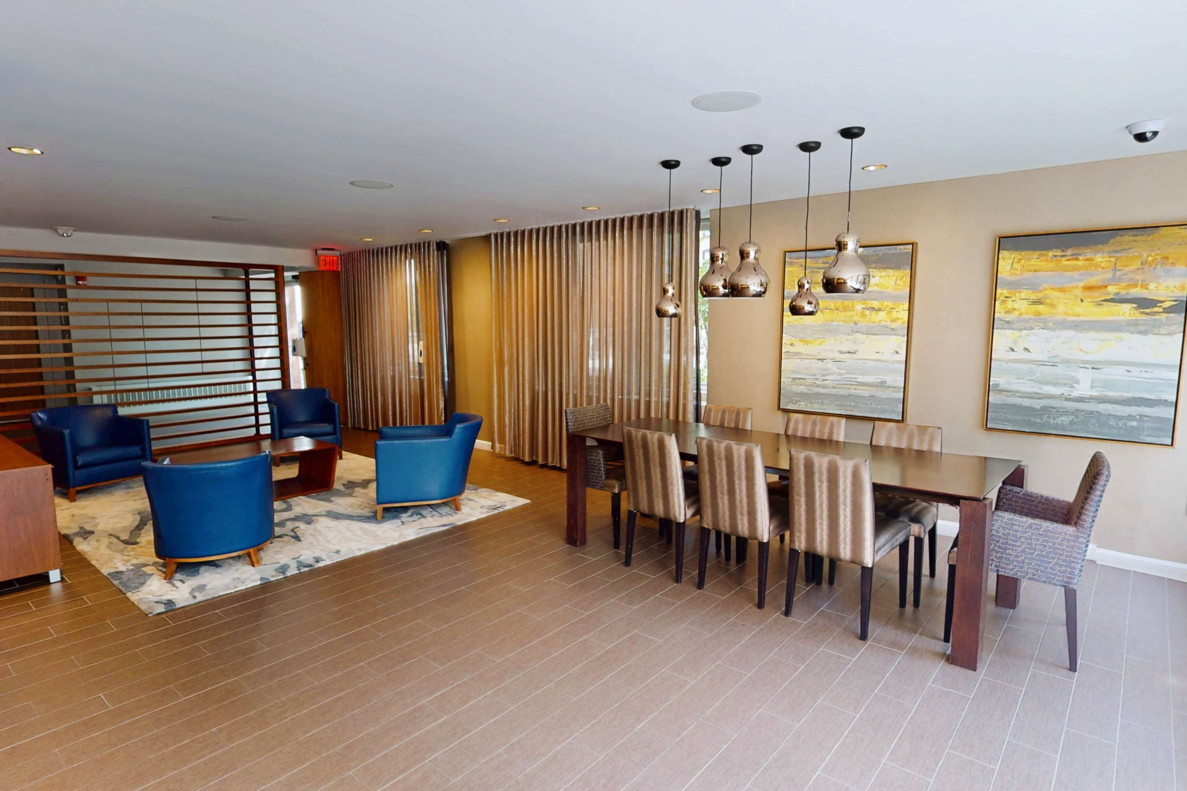 The image shows a modern lounge area with two blue chairs, a marble-patterned rug, a dining table with eight chairs, and two large paintings on the walls.