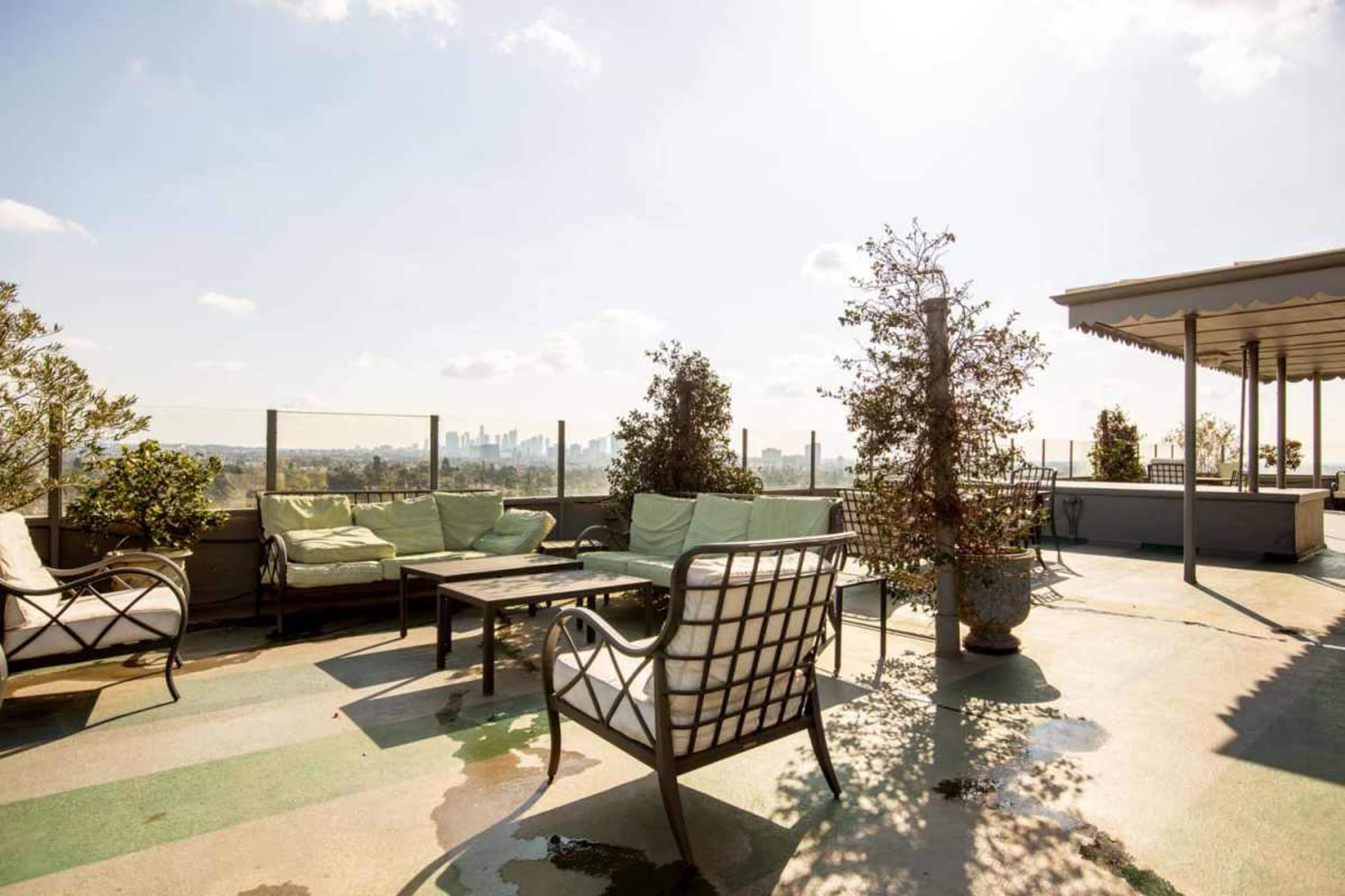 The rooftop features several outdoor seating arrangements, including chairs and a table, surrounded by potted plants under a clear sky.
