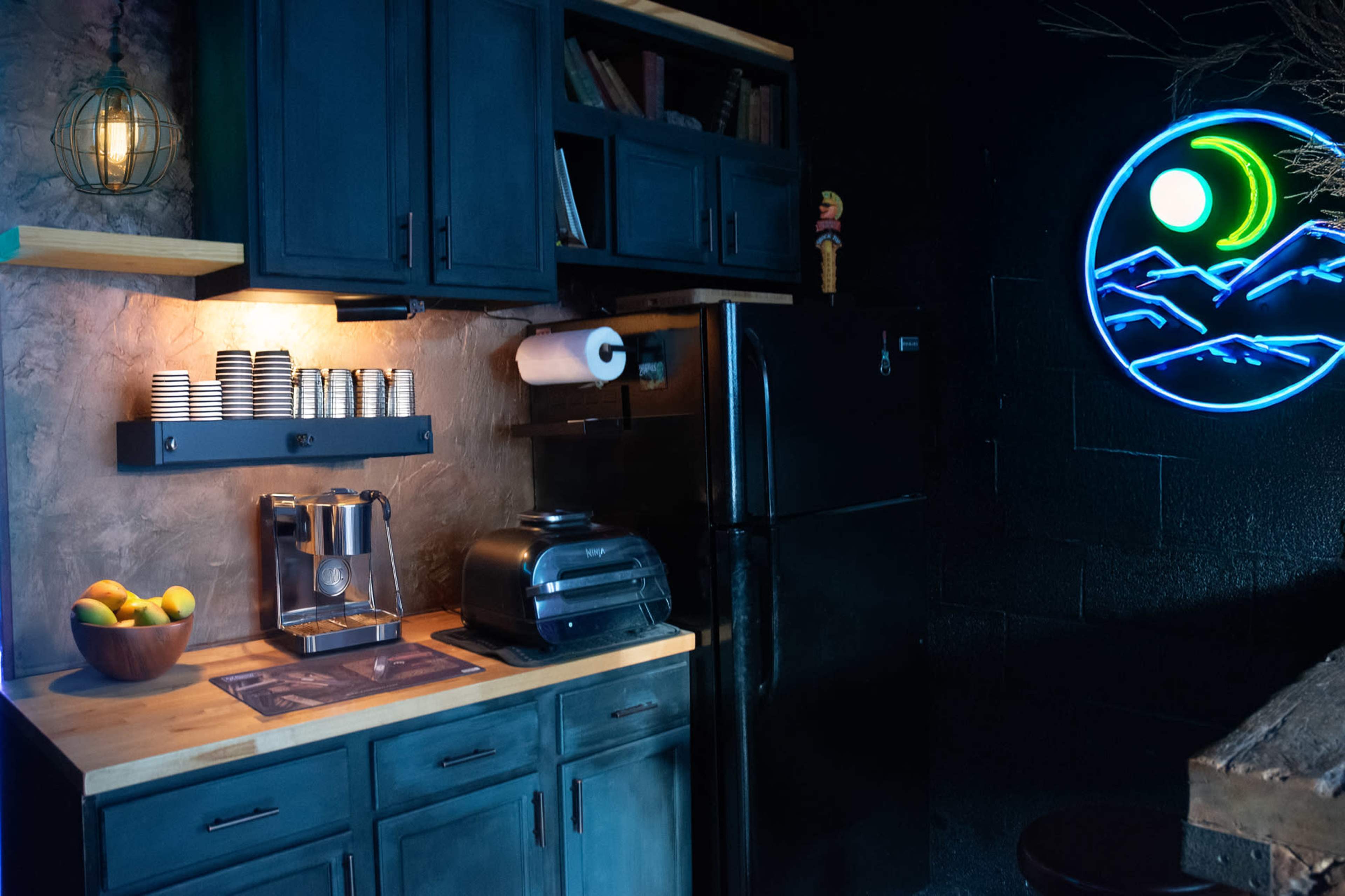 The image shows a dark-themed kitchen area with a wooden countertop, a coffee maker, a toaster, a bowl of fruit, and a neon sign depicting mountains and a crescent moon.