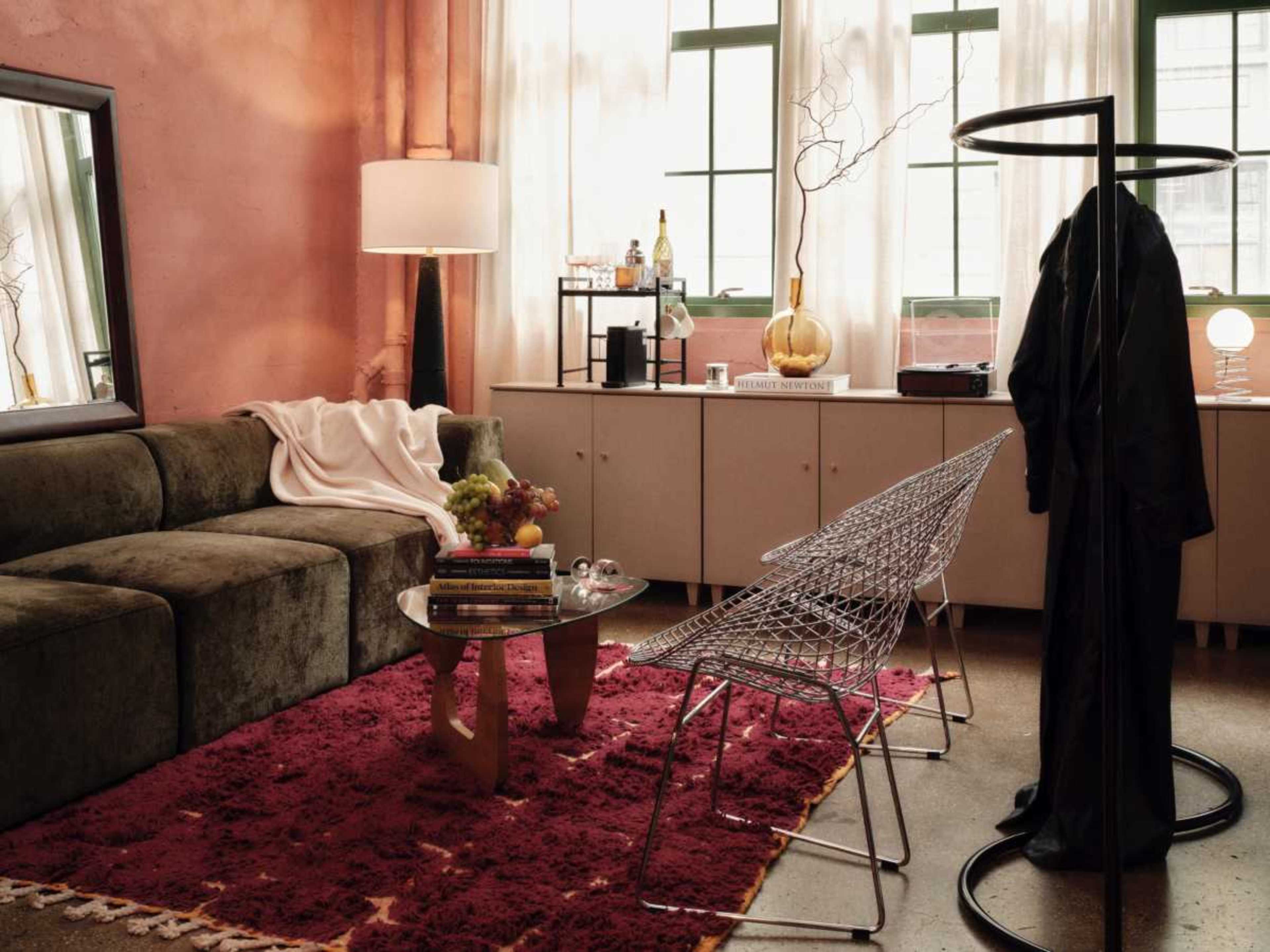 The image shows a cozy living room featuring a green sofa, a circular coffee table with books, two wire chairs, and a side table with decorative items, set against a backdrop of pink walls and large windows.