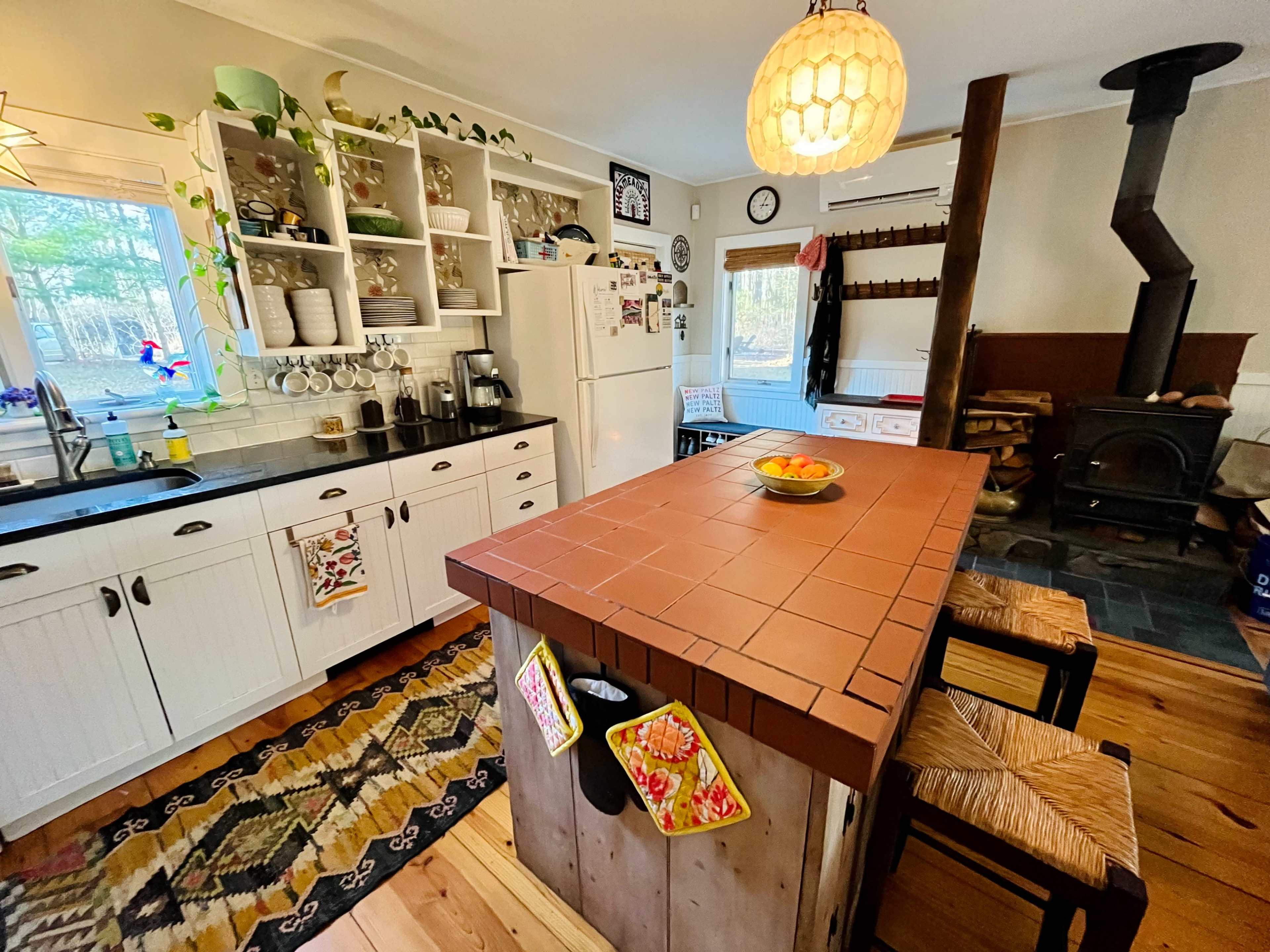 The kitchen features a central island with a tiled top, surrounded by wooden stools, and is equipped with white cabinets and a cozy wood-burning stove in the corner.