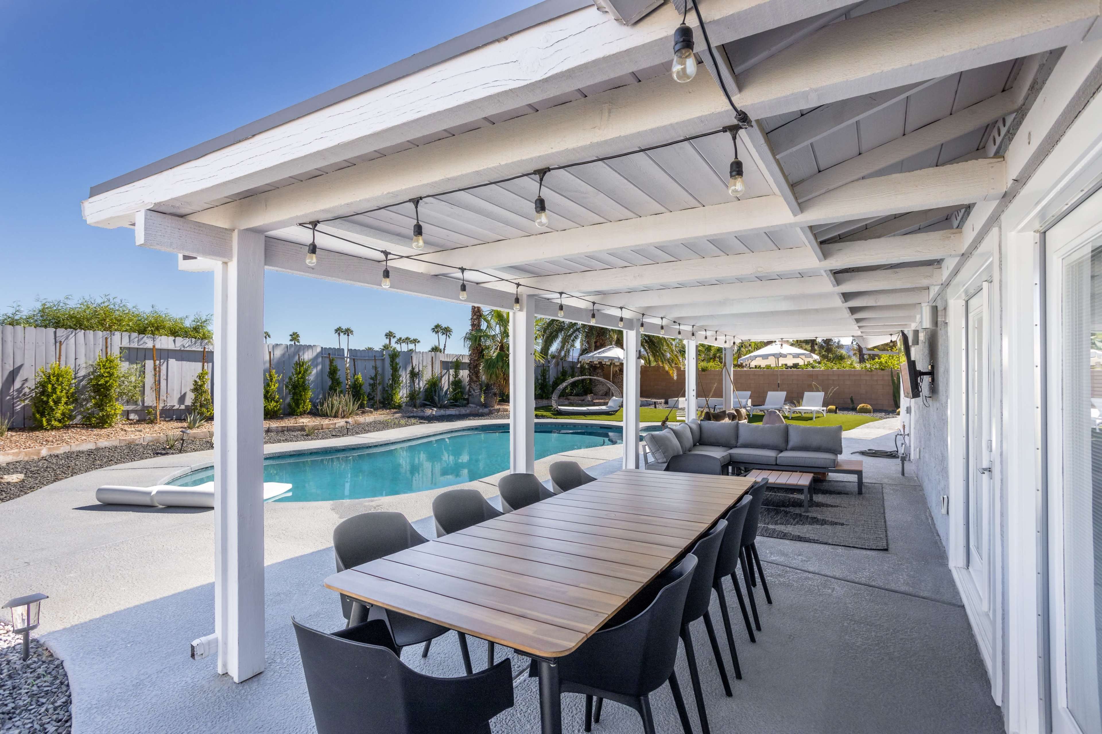 A covered patio with a long dining table and chairs overlooks a swimming pool surrounded by landscaping.
