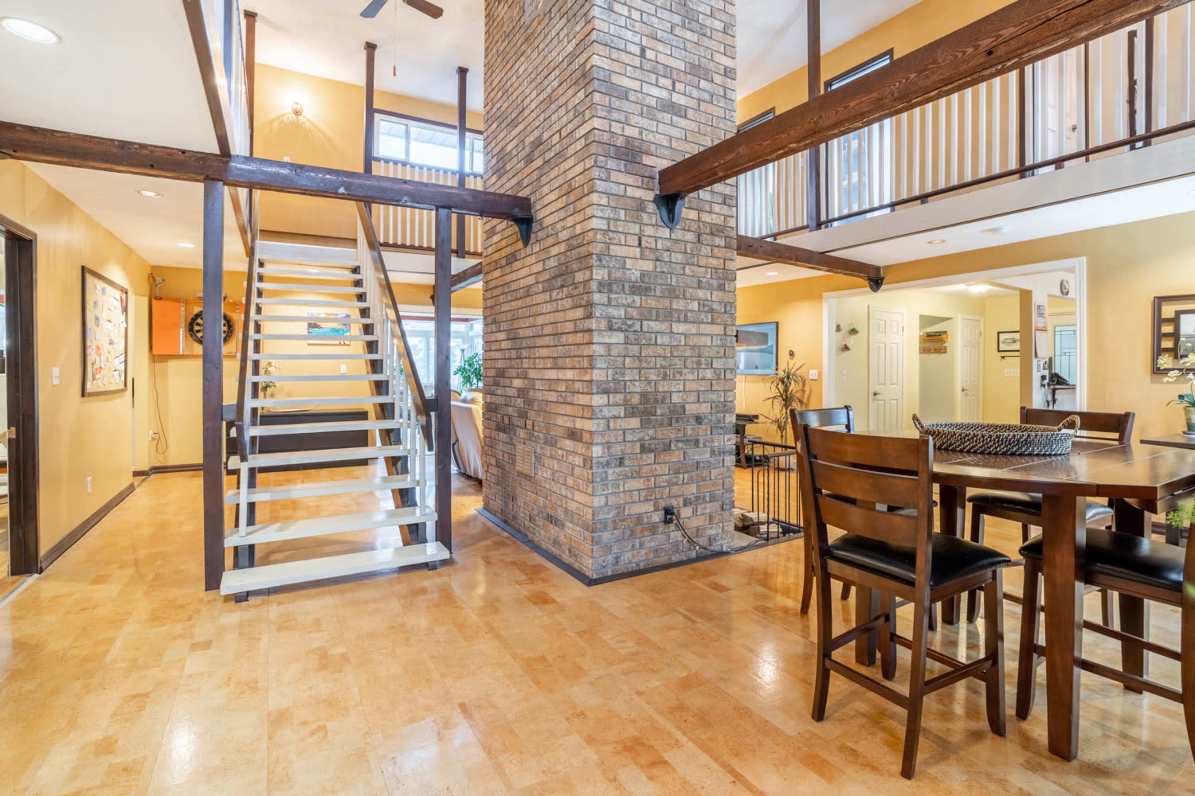 The image shows a spacious interior with a central brick column, a staircase leading to an upper level, and a dining area with wooden furniture.